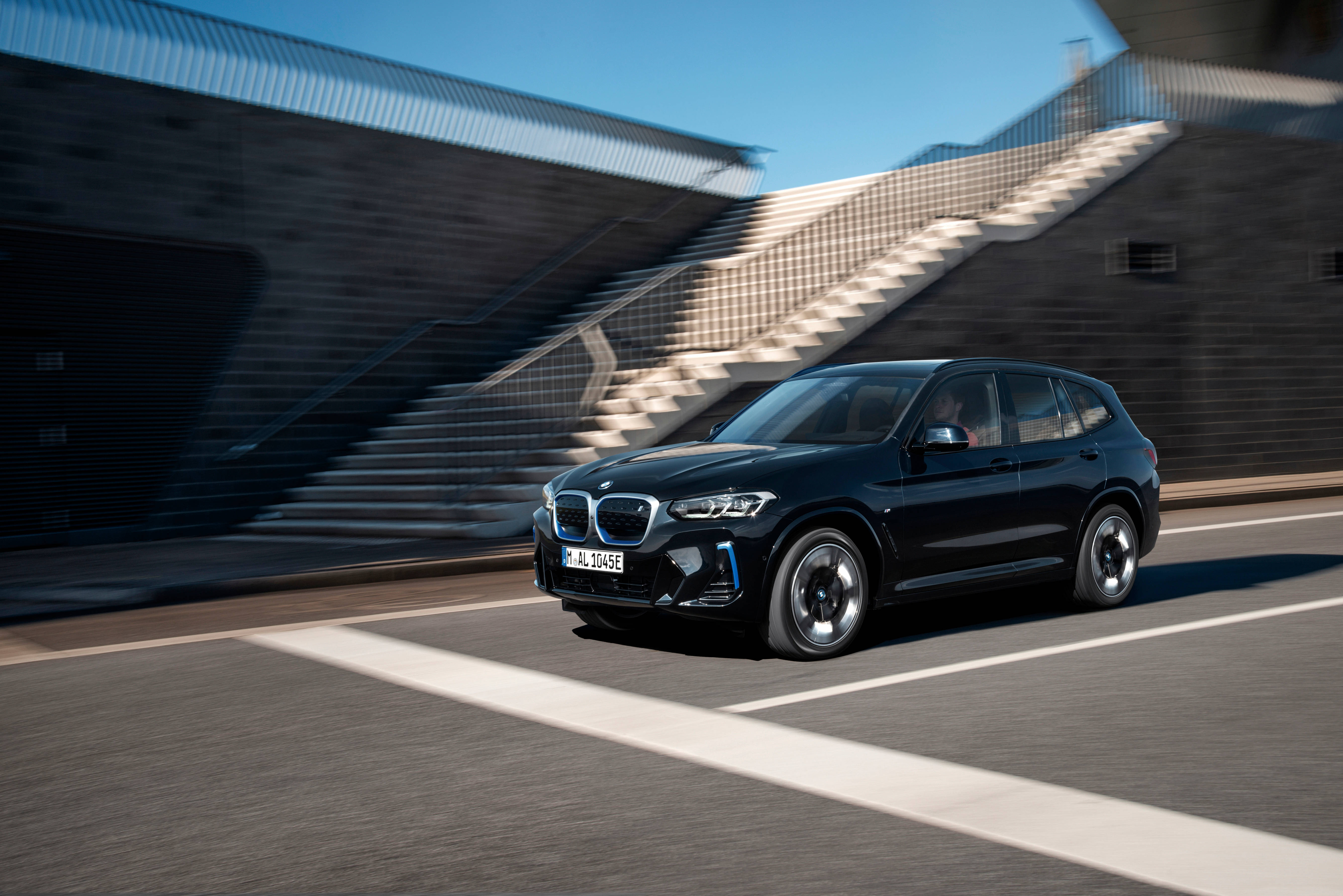 黑色BMW IX3 M Sport汽車 壁紙 — 一架時尚黑色BMW SUV在現代城市道路上疾馳，帶有動態模糊效果，背景是幾何混凝土樓梯和晴朗藍天。