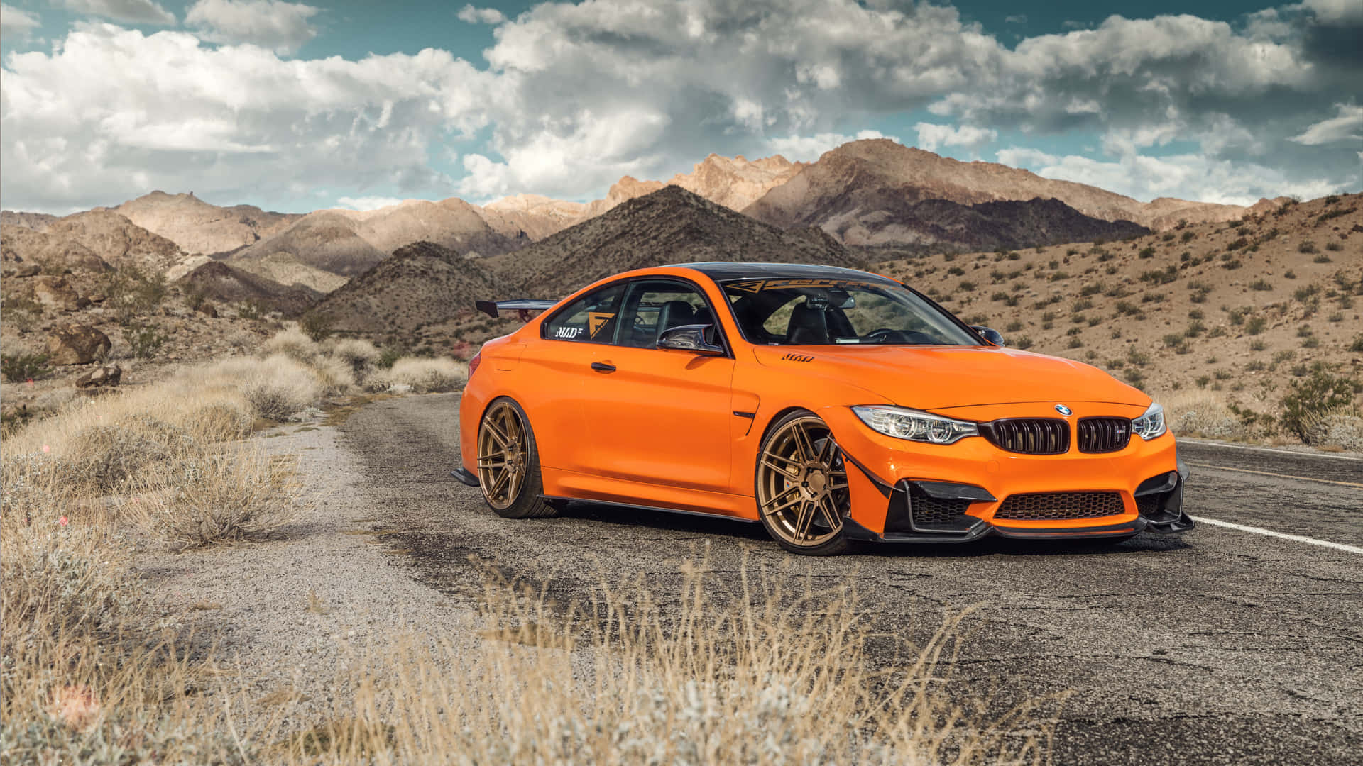 駕馭風格與豪華：駕駛BMW 壁紙 — 一輛亮橙色BMW M4雙門跑車配金色輪圈，停泊在沙漠公路上，背景是崎嶇山脈和陰天。