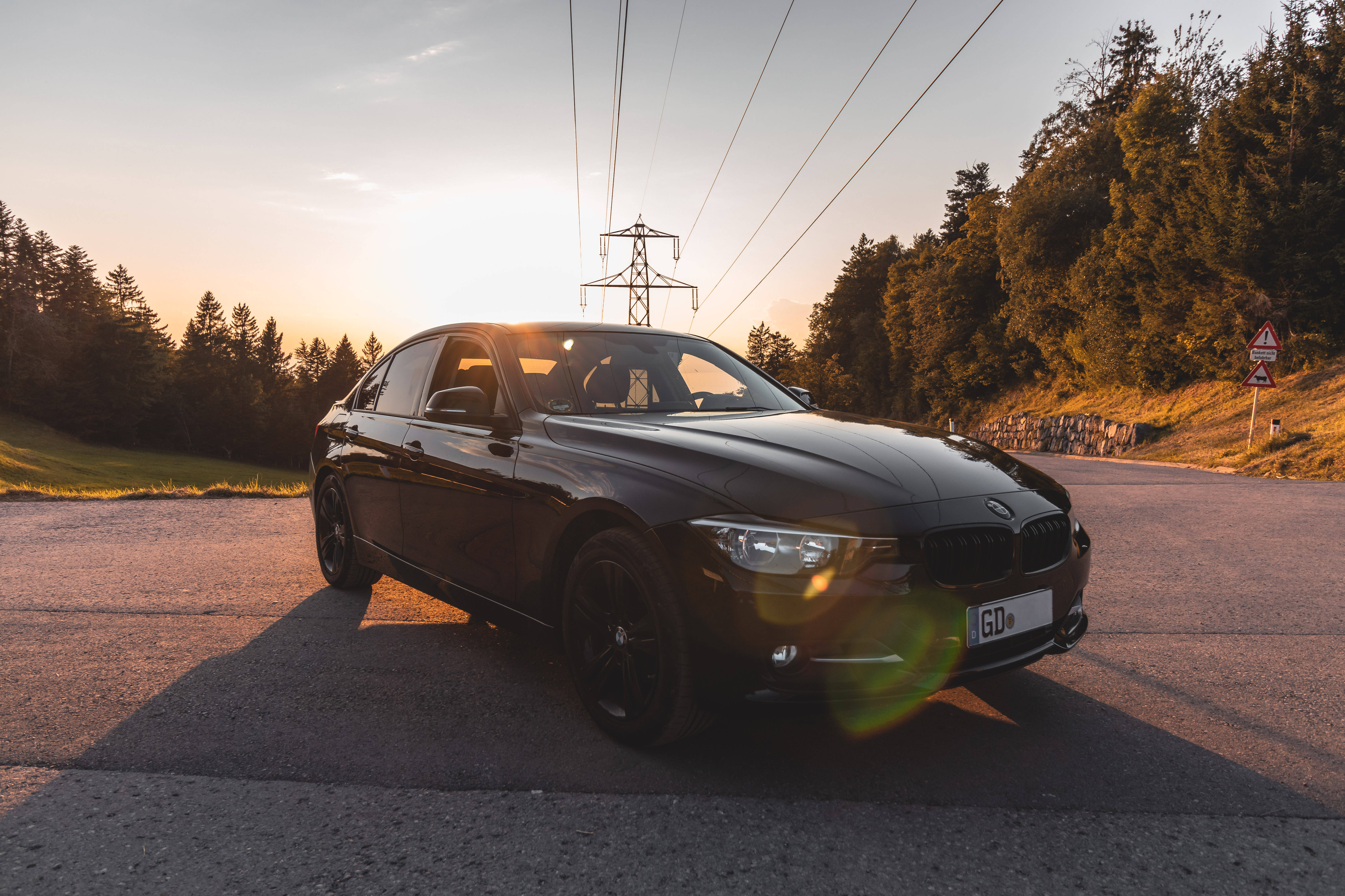 黑色BMW M系列於森林中 壁紙 — 一輛時尚的黑色BMW房車停泊在蜿蜒的山路上，背景是金色的日落、電線和森林。