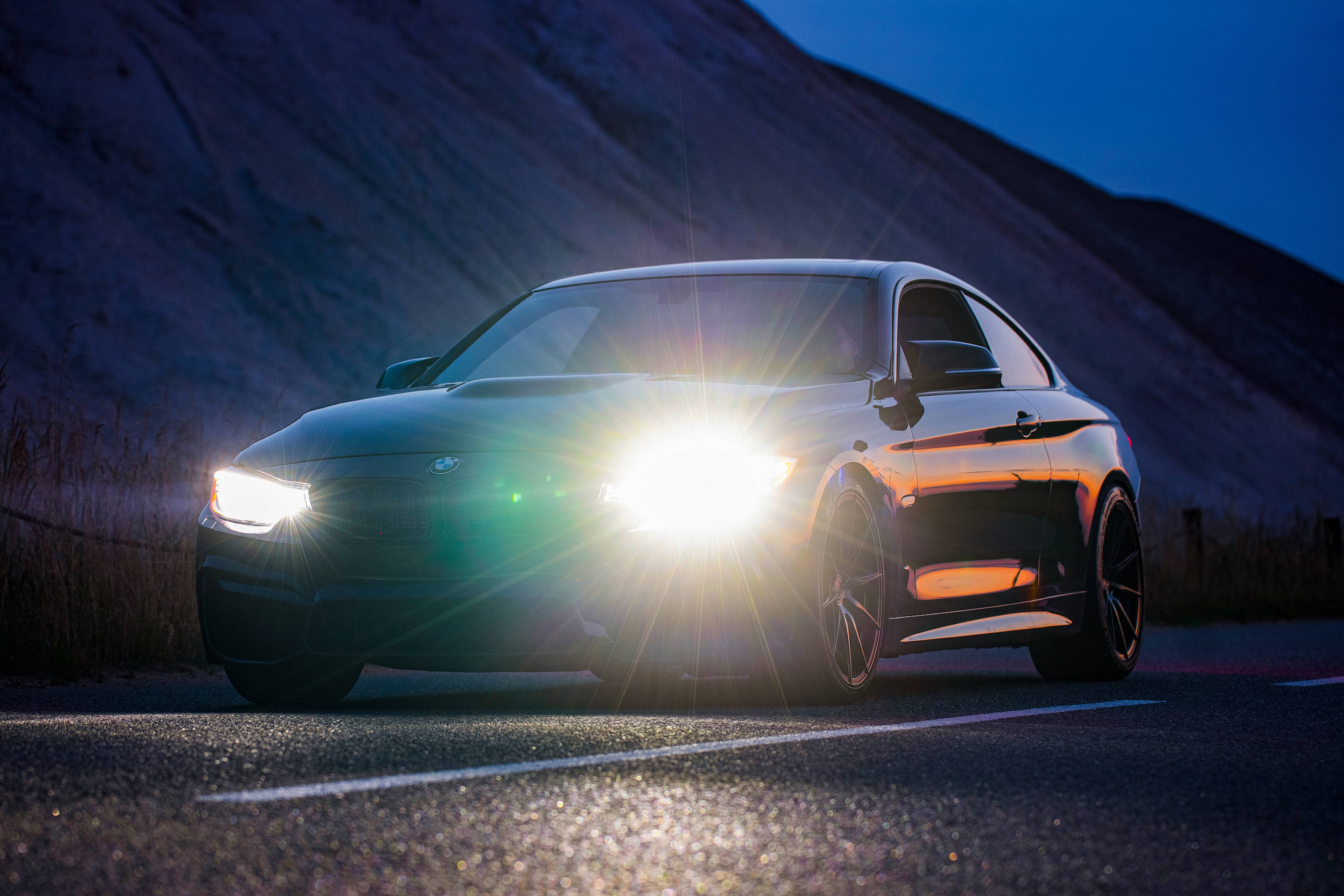 夜間路中BMW M頭燈亮起 壁紙 — 一輛時尚的黑色跑車，配備發光的車頭燈，在山路上劃破暮色，在濕漉漉的瀝青上投射出銳利的倒影。