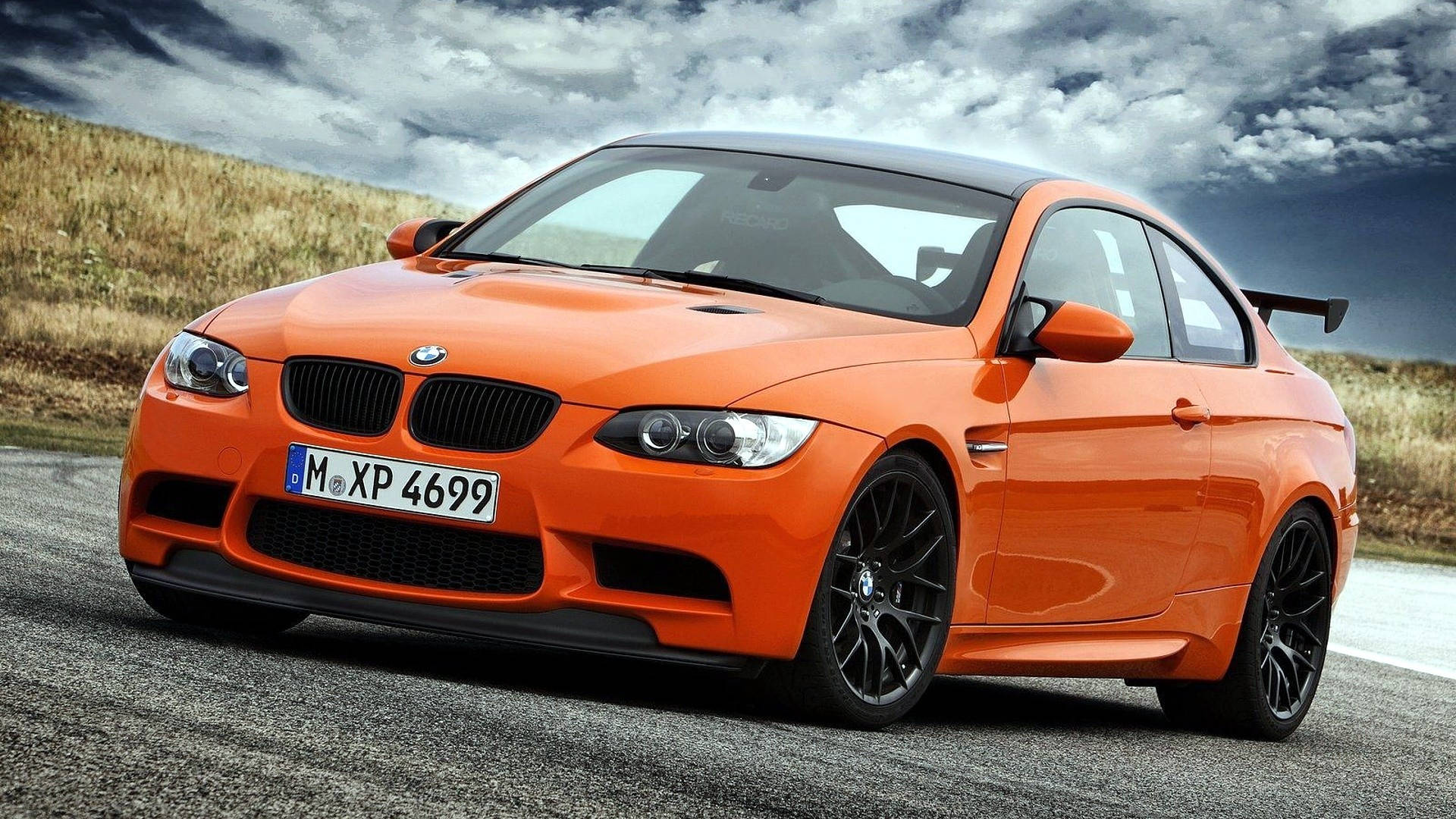 橙色BMW M車 壁紙 — 一輛充滿活力的橙色BMW M3雙門轎跑車，帶有黑色飾件和後擾流板，停在碎石路上，天空烏雲密佈，氣氛戲劇化。