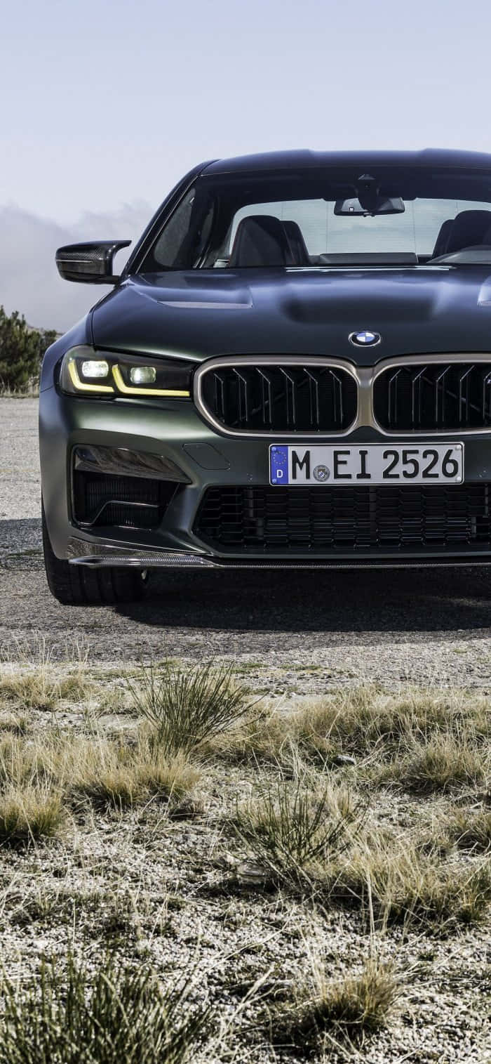 於 BMW M 中享受 iPhone 的豪華體驗 壁紙 — 一架時尚的啞光黑色BMW M5停泊在碎石上，乾草叢生，天空晴朗。