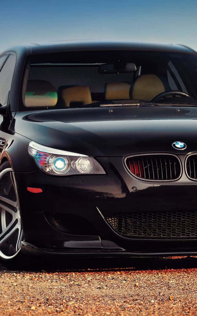 黑色 BMW M 車旁置放 iPhone 壁紙 — 一輛時尚的黑色寶馬跑車，車頭燈閃爍，停在碎石地上，背景是晴朗的藍天。