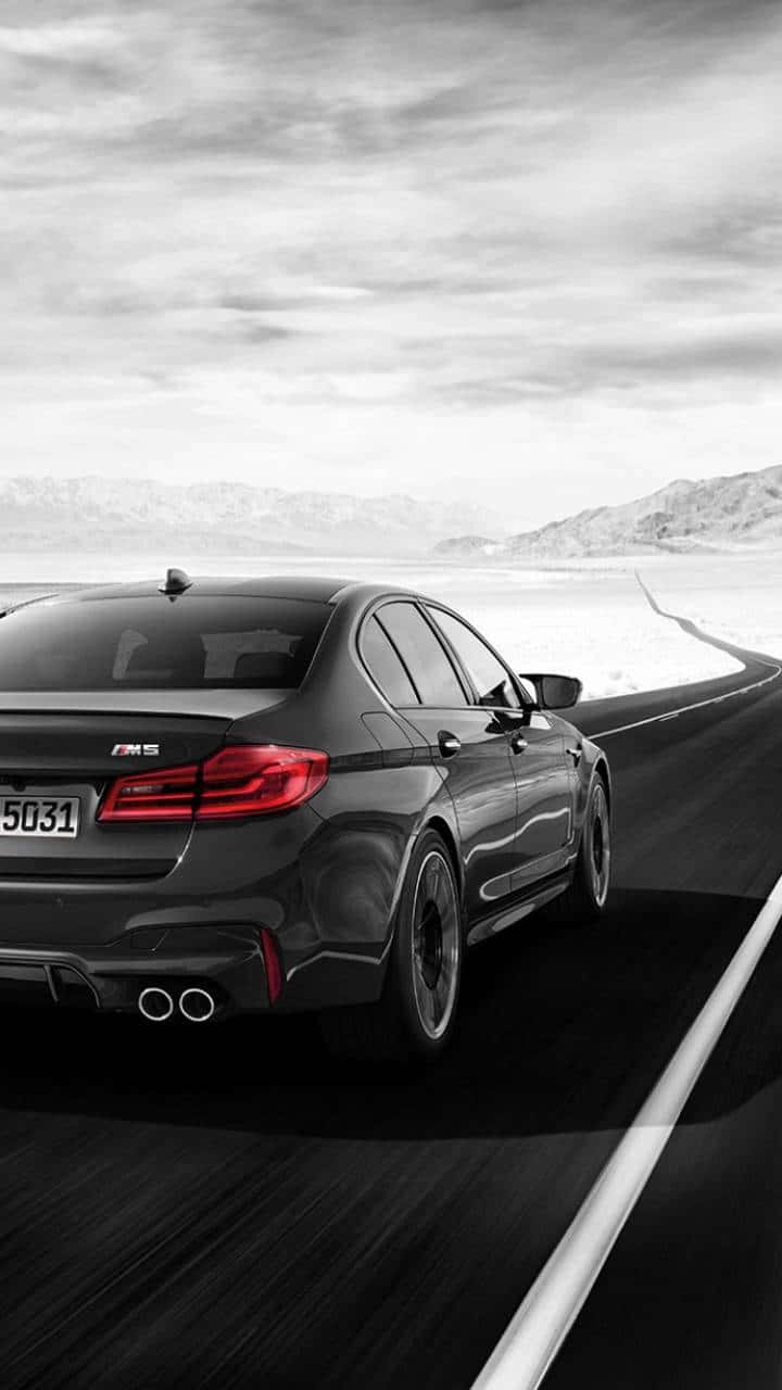 BMW M 系列於 iPhone 上呈現 壁紙 — 一輛時尚的黑色BMW M5在陰天、遠山背景下，沿著蜿蜒的沙漠公路疾馳。