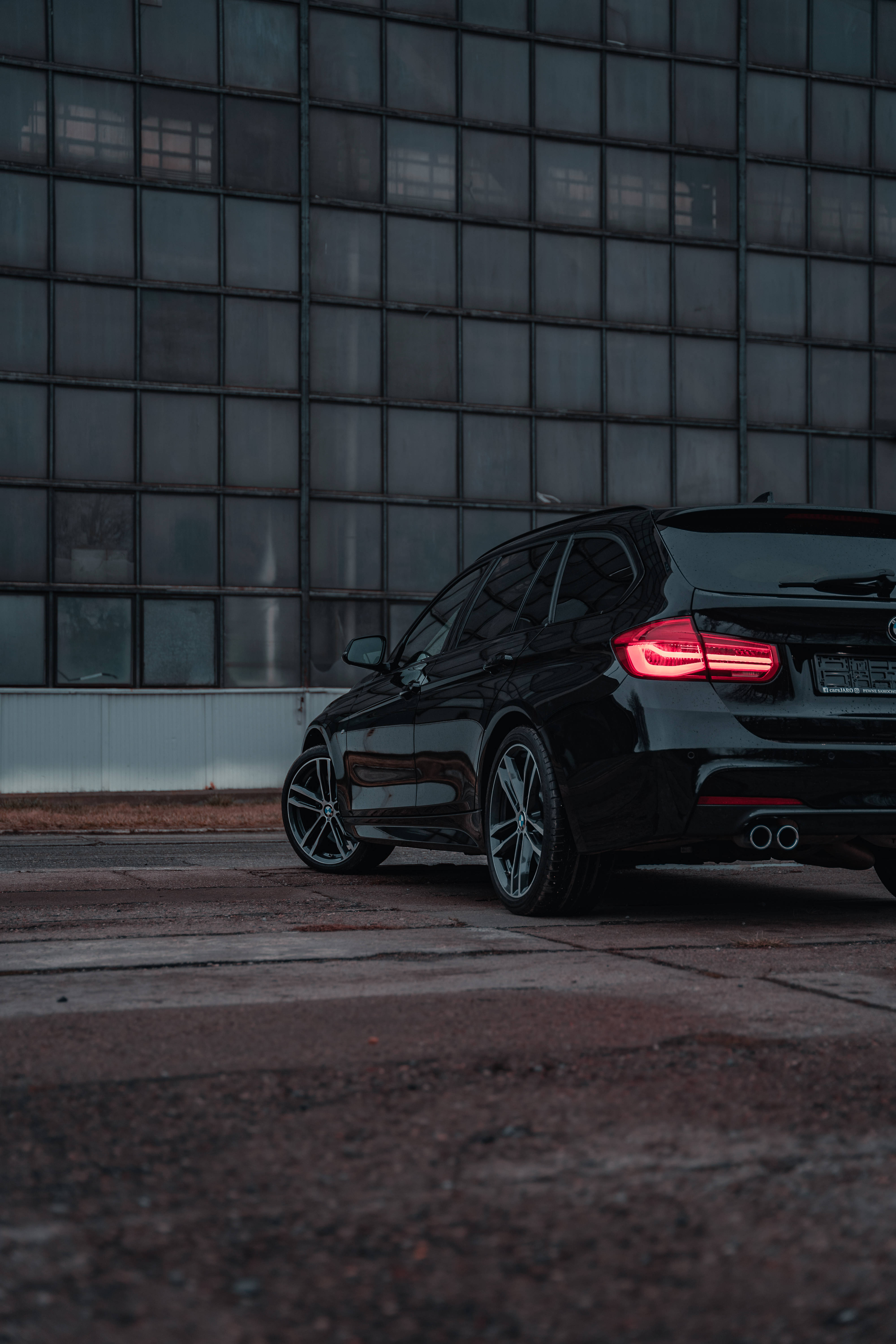 標題：運動美學 — BMW M系列車停泊於建築藝術交融之處 壁紙 — 一輛時尚的黑色BMW旅行車在黃昏時停泊在網格狀玻璃建築前，紅色尾燈閃爍發光。