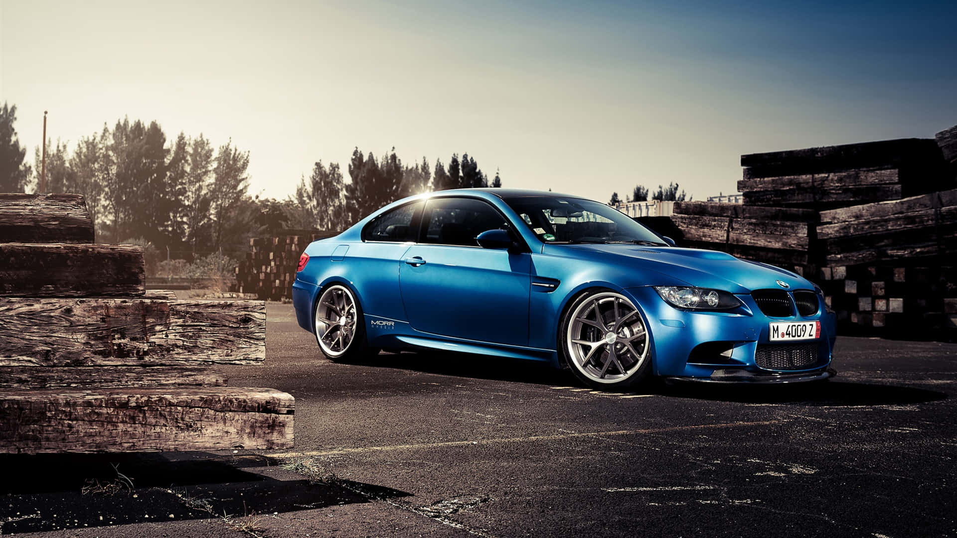 BMW M Performance 壁紙 — 一架充滿活力的藍色BMW M3雙門跑車停在瀝青路上，旁邊是堆疊的風化木樑，天空晴朗。