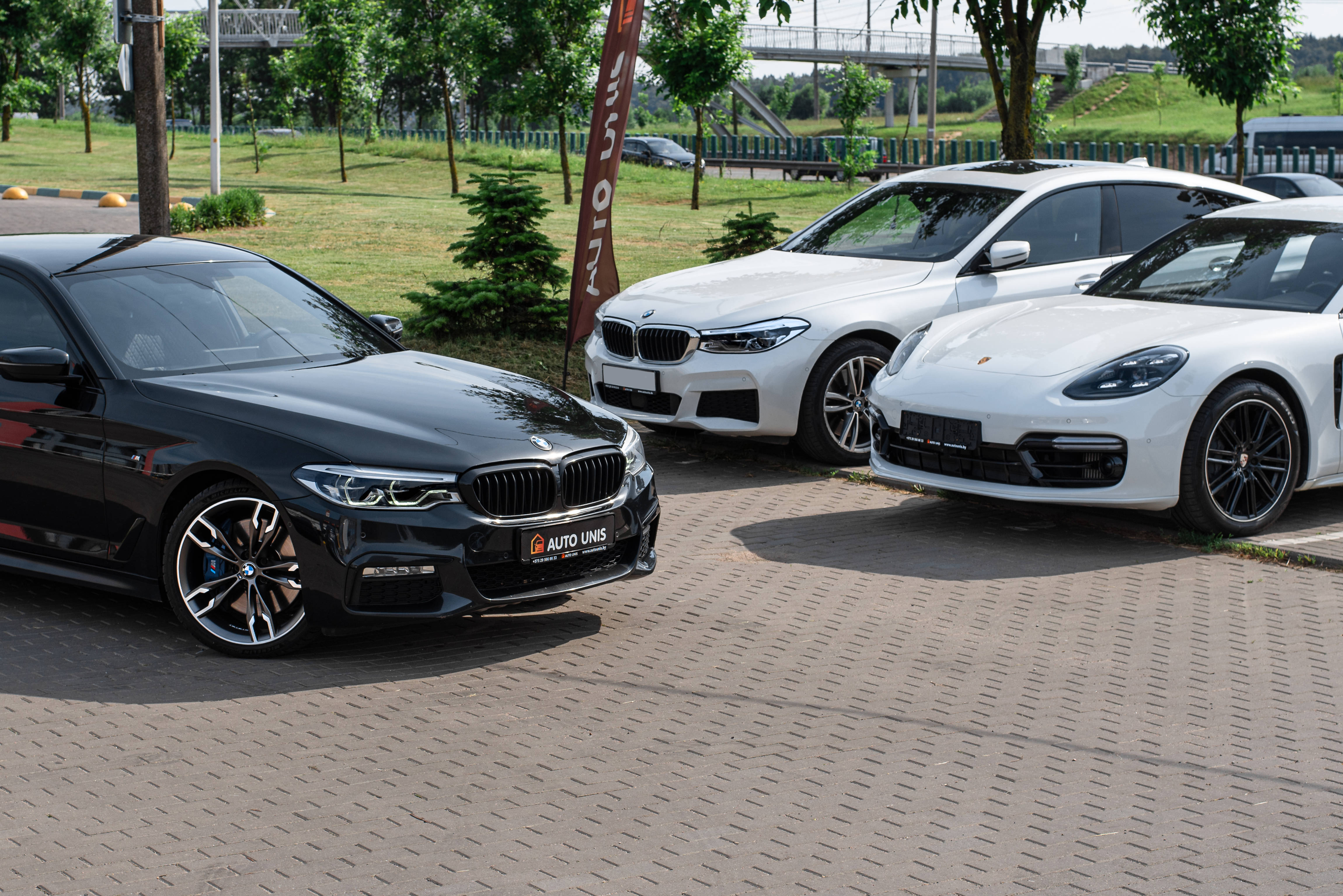 BMW M系列於車展現場 壁紙 — 三輛豪華車 黑色BMW轎車 白色BMW轎車 白色保時捷Panamera 停放在鋪砌的停車場 背景有綠樹和橋樑