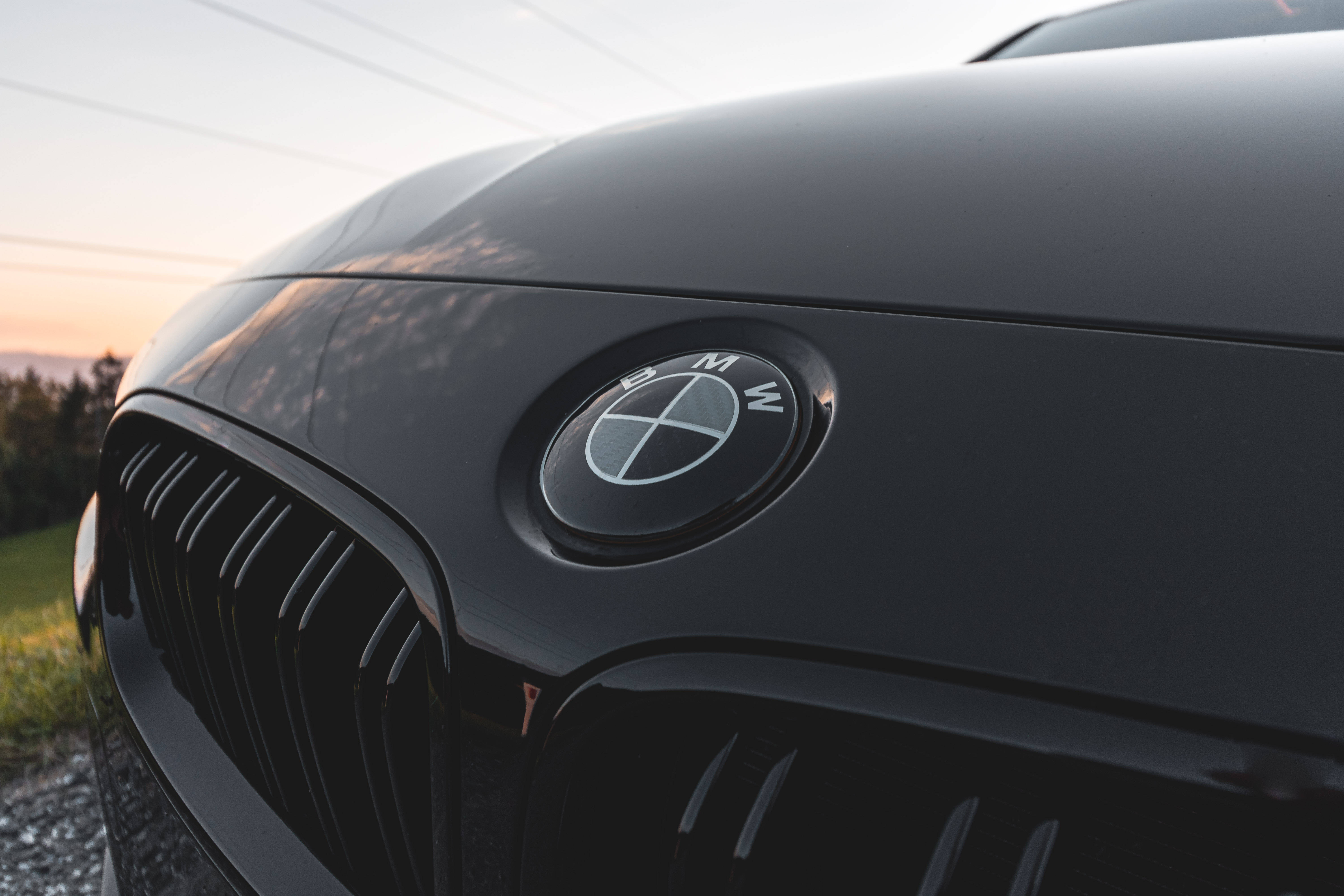 BMW M系列前標誌 壁紙 — 日落時分，溫暖天空倒影和森林背景下，時尚黑色BMW引擎蓋的特寫。