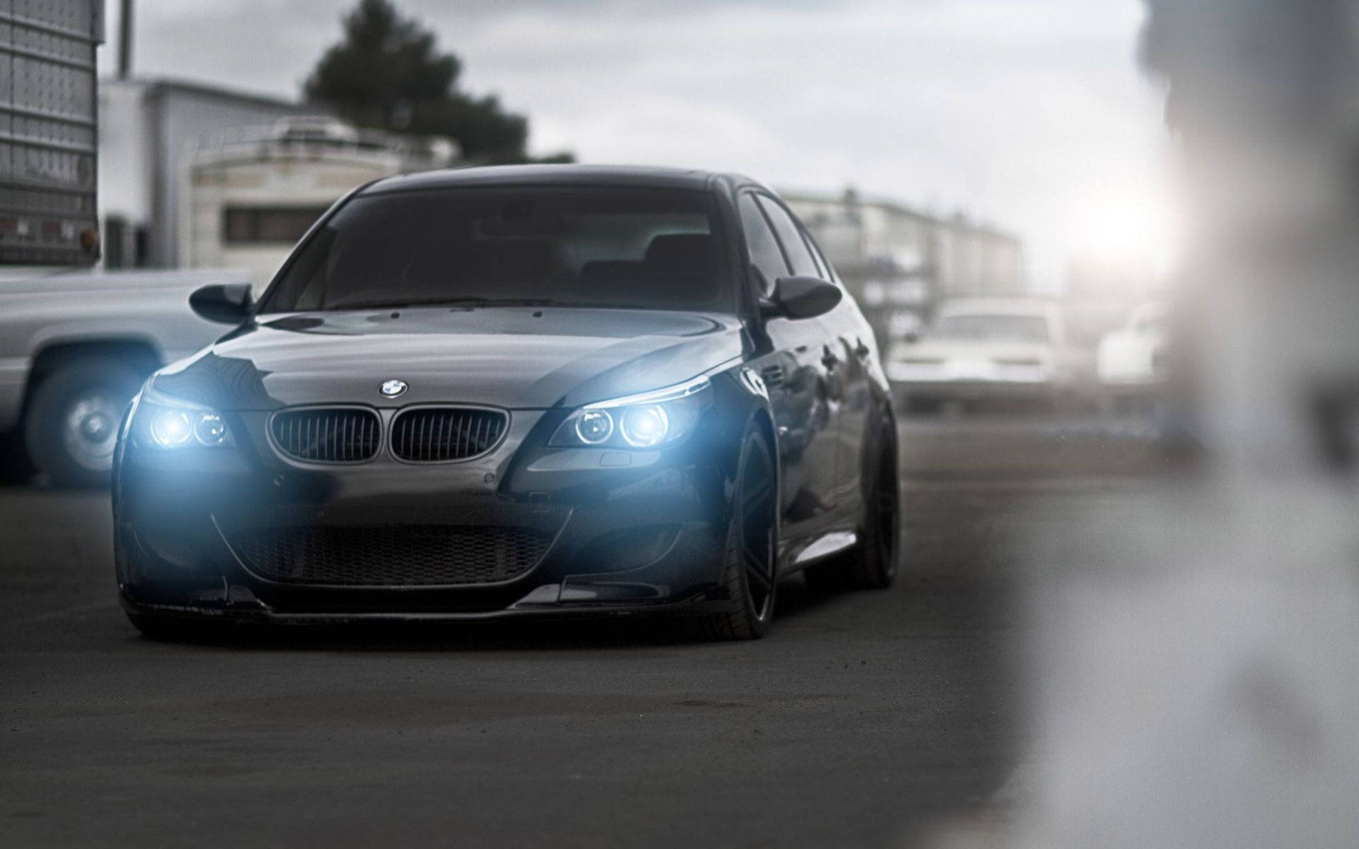 BMW M 系列攝影 壁紙 — 一輛時尚的黑色BMW M5跑車，配備發光的車頭燈，在多雲的天空下主宰著一個粗糙的城市地段。