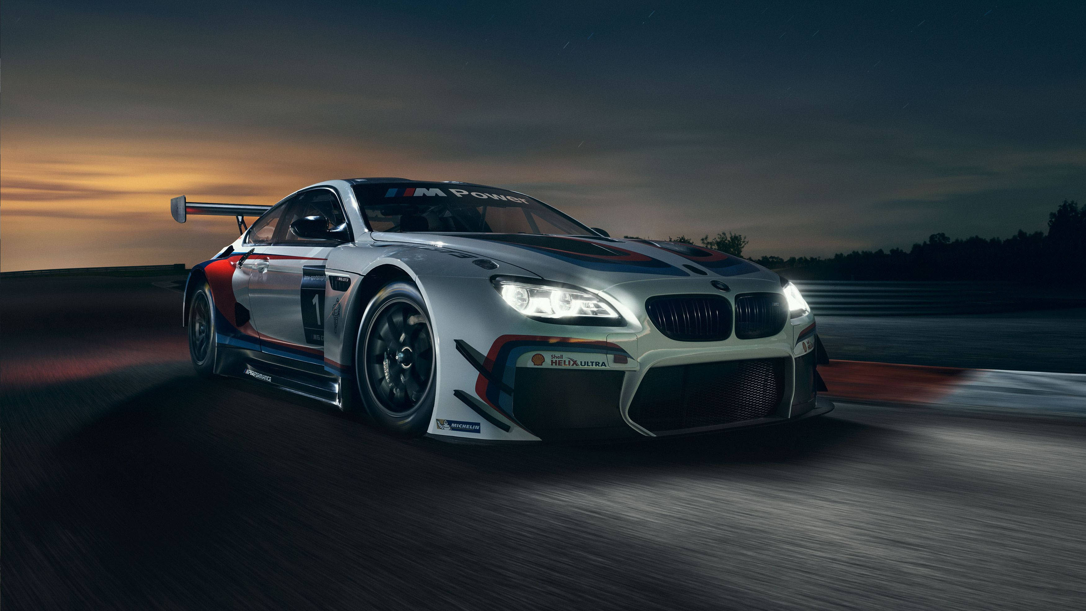 BMW M 系列賽車 壁紙 — 一輛銀色BMW M6 GT3賽車在黃昏時分沿著賽道疾馳，車頭燈閃爍，帶有動態模糊效果。