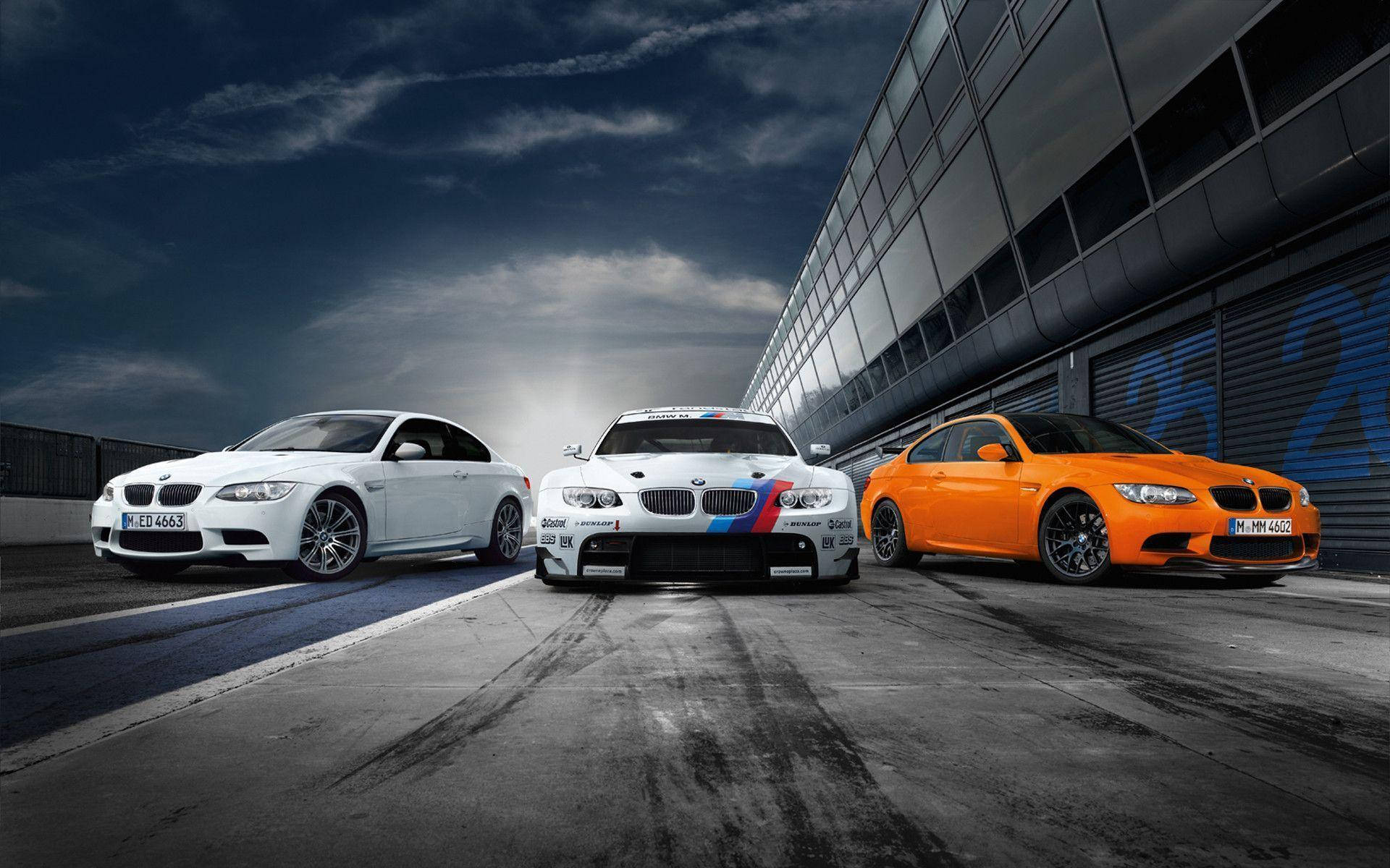 BMW M 系列車型 壁紙 — 三輛寶馬跑車，白色、橙色和白色，在暴風雨的天空下，在現代建築旁邊的潮濕賽道上排成一排。