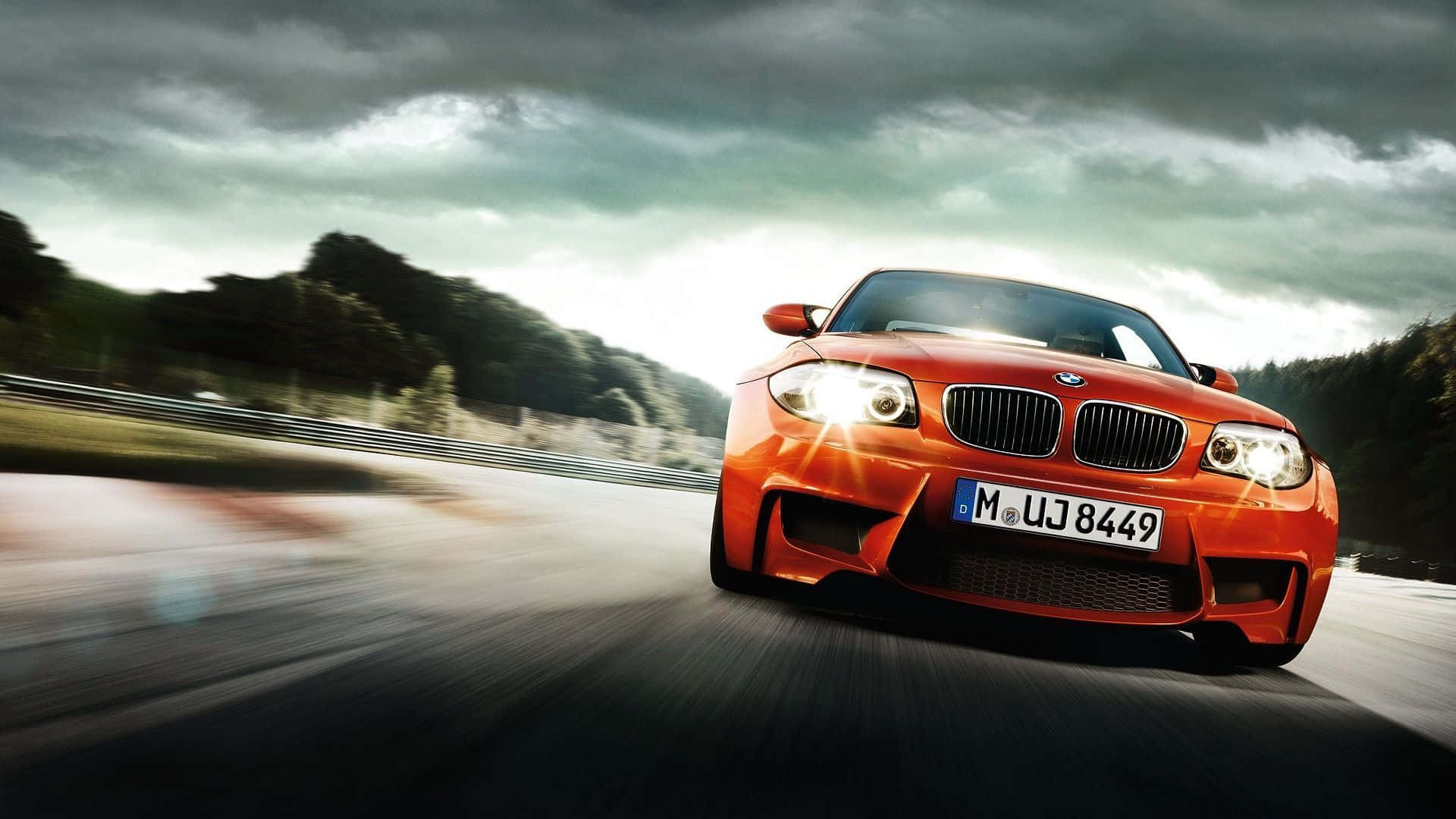 BMW 桌面橘色 M1 系列 壁紙 — 一輛充滿活力的橙色BMW M1在暴風雨的天空下，在潮濕的道路上高速行駛，帶有動態模糊效果。