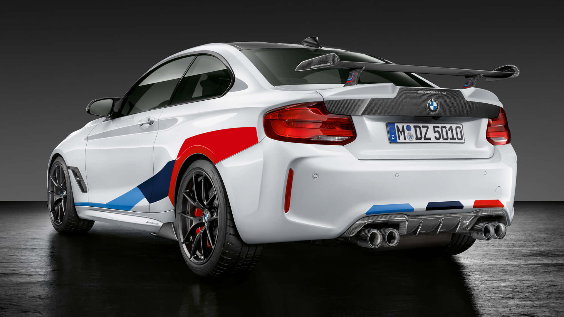 白色 BMW M2 1920x1080 M 系列背景 壁紙 — 一輛白色BMW M2雙門轎跑車，帶有紅藍賽車條紋和大型後擾流板，在黑暗的攝影棚中以左後方四分之三角度展示。