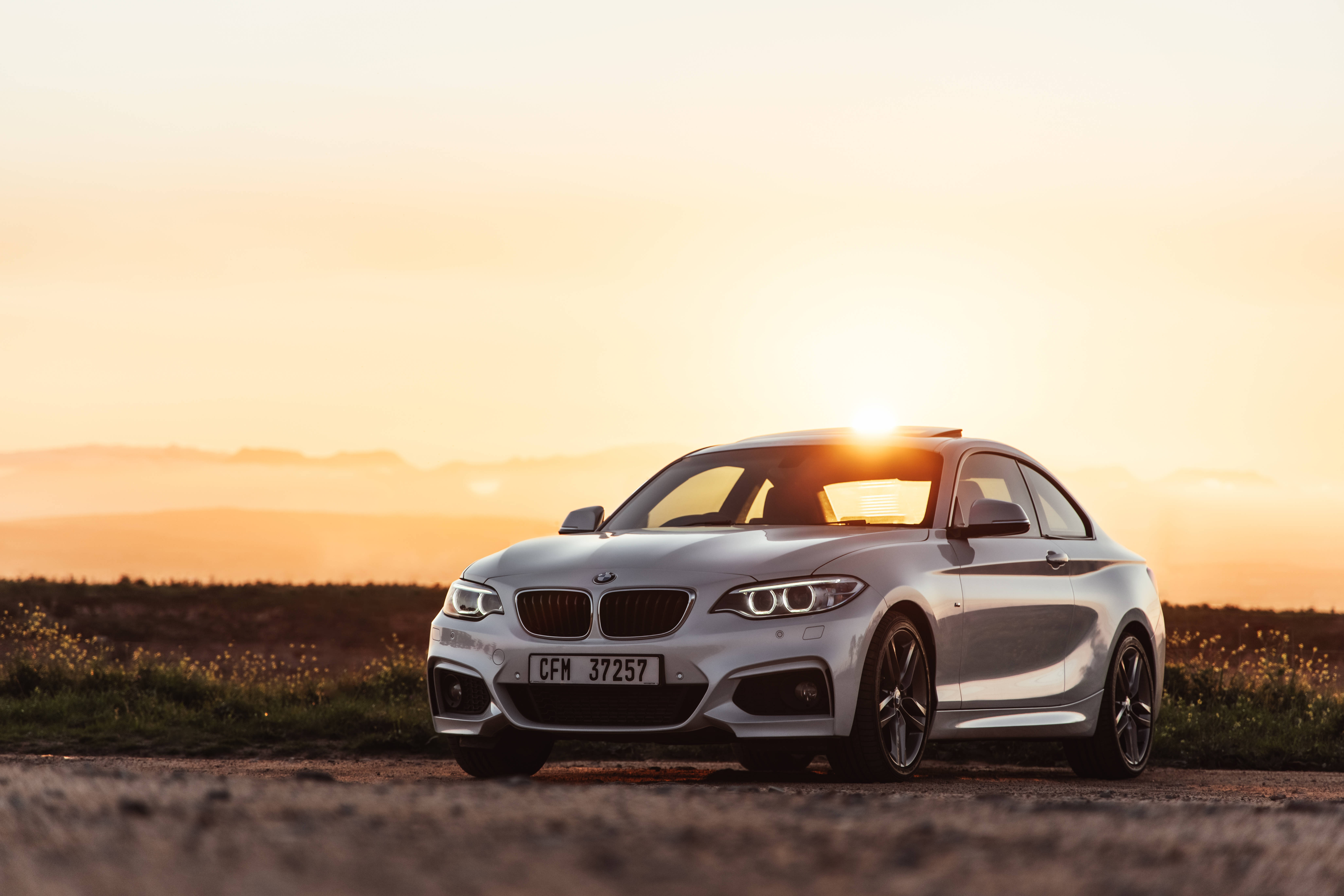 BMW M2系列跑車 壁紙 — 一輛銀色寶馬轎跑車在金色日落下的鄉村泥路上閃閃發光，遠處有山丘。