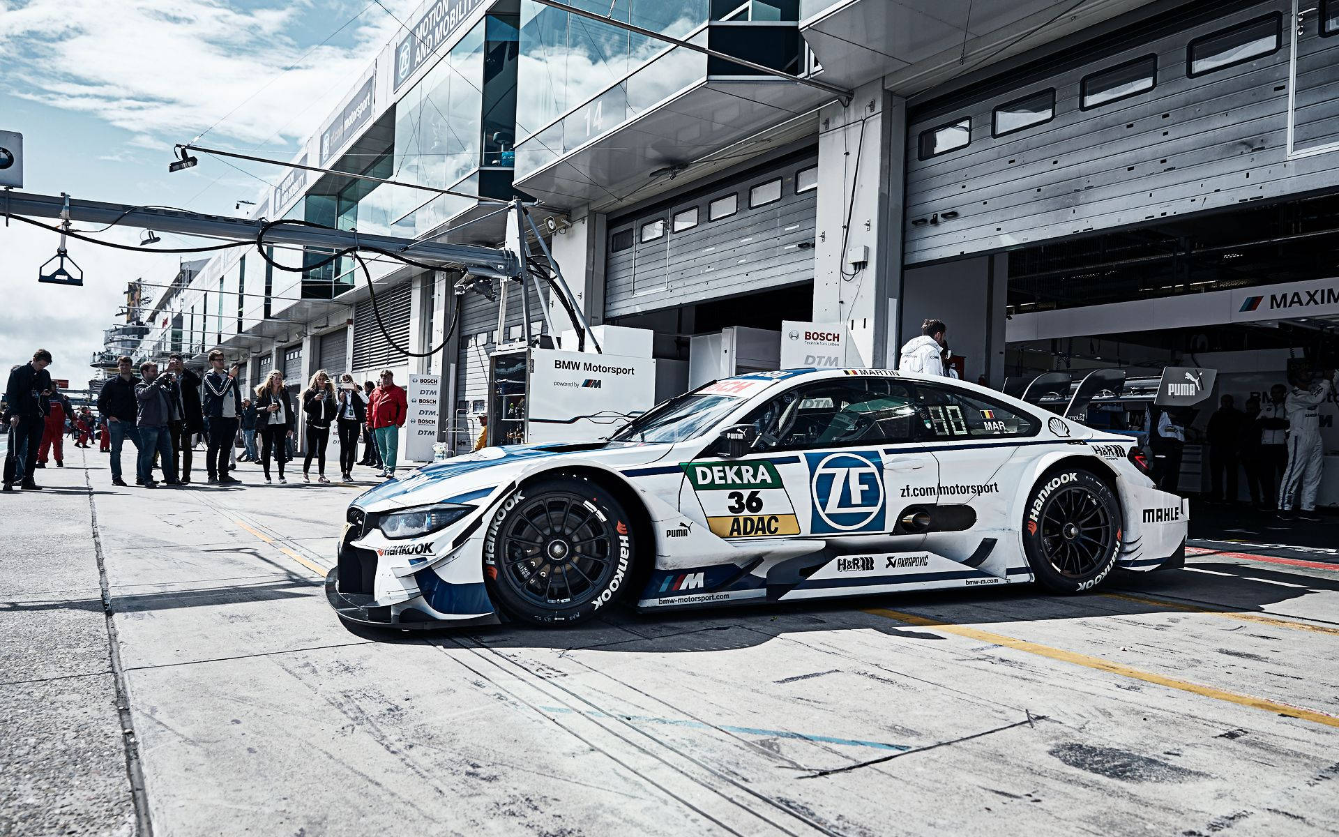 BMW M3 DTM賽車系列 壁紙 — 一輛帶有藍色和黑色點綴的白色BMW賽車，停在賽車活動的維修區車庫內，天空部分多雲。