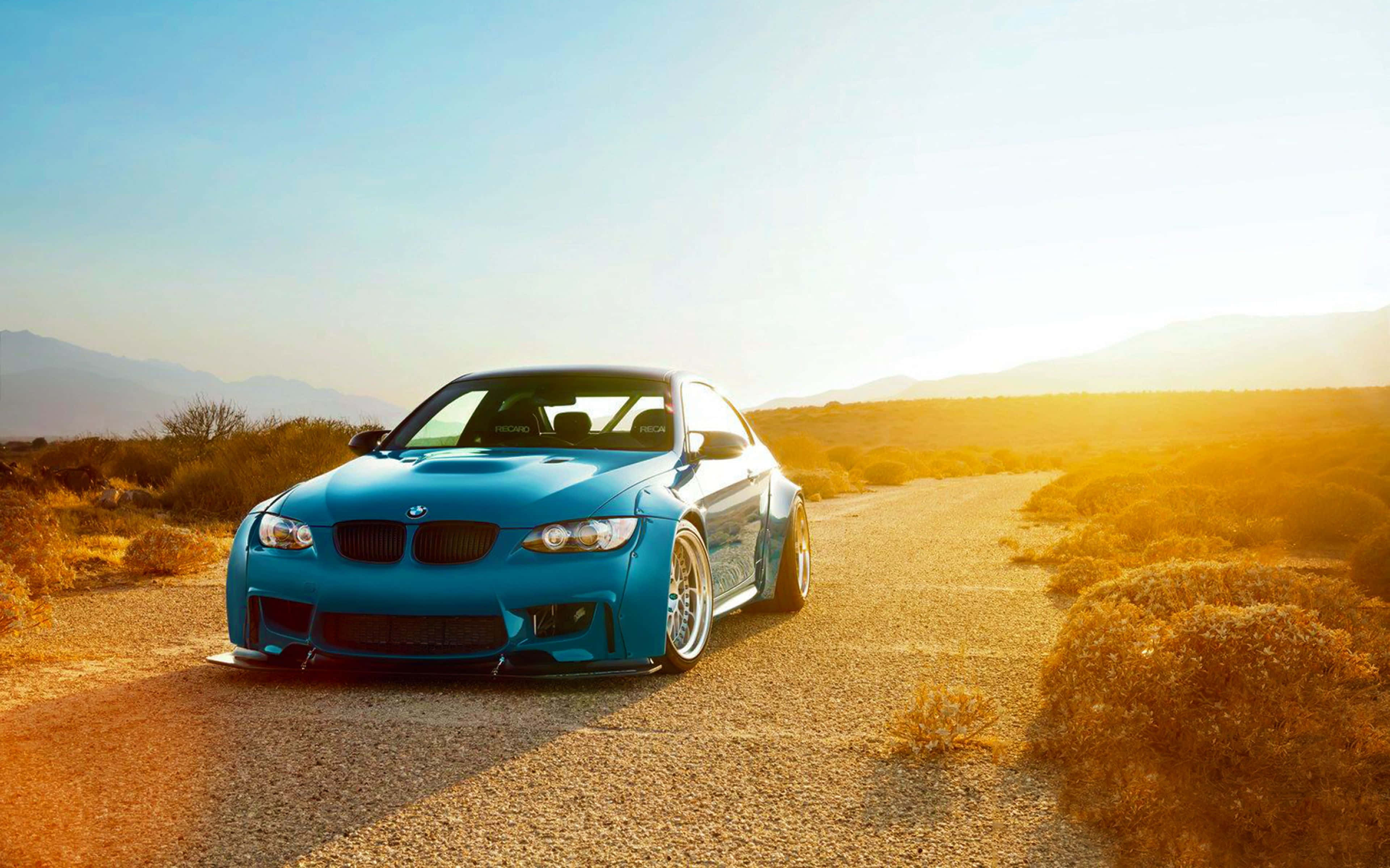 BMW M3客製化平板壁紙 壁紙 — 一輛鮮豔的藍色BMW跑車停泊在陽光明媚的沙漠公路上，周圍是金色的山丘和遠處的山脈，天空晴朗。