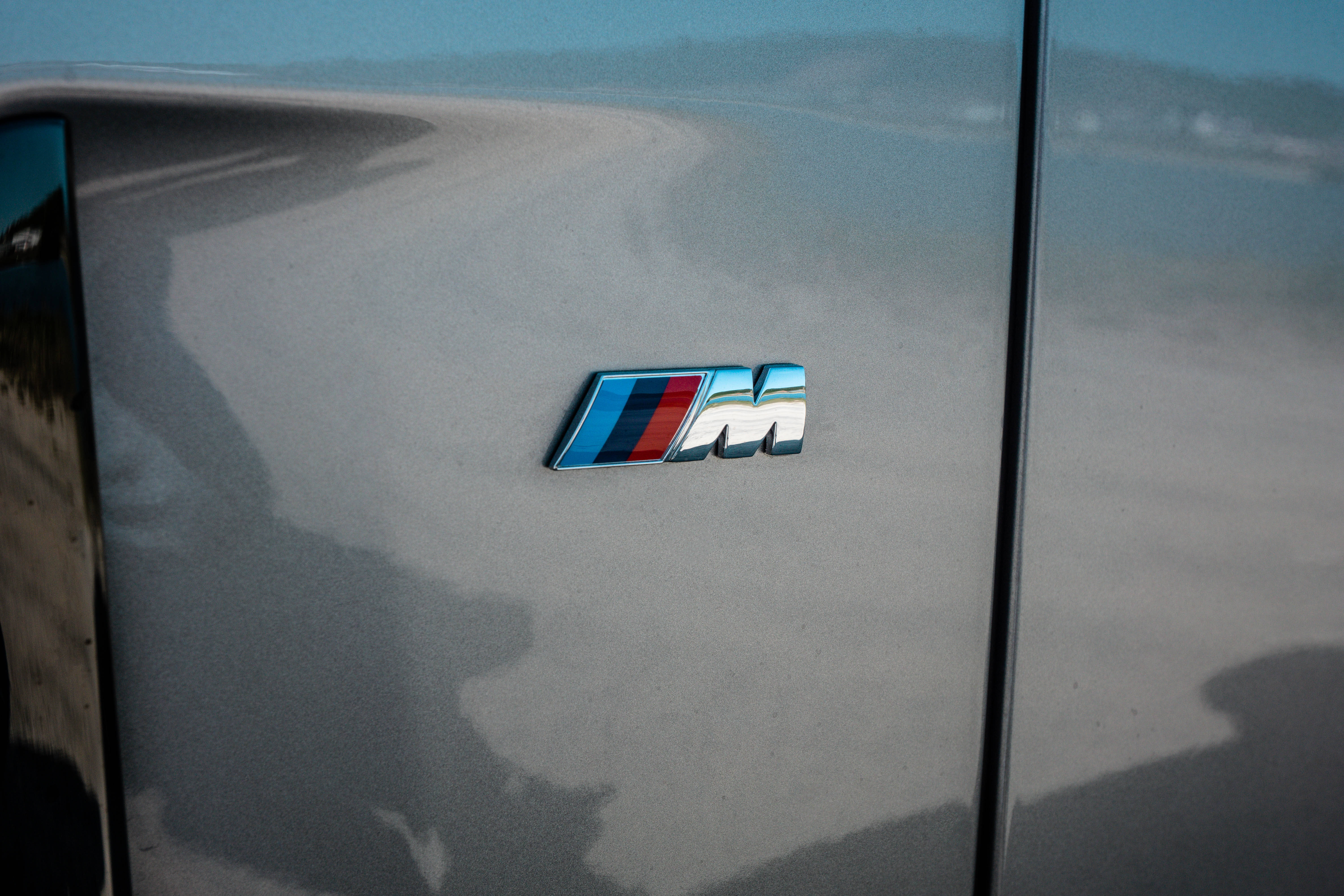 BMW M3標誌徽章特寫 壁紙 — BMW M 徽章特寫，位於時尚車門上，反射出海岸道路和遠山，在柔和日光下。