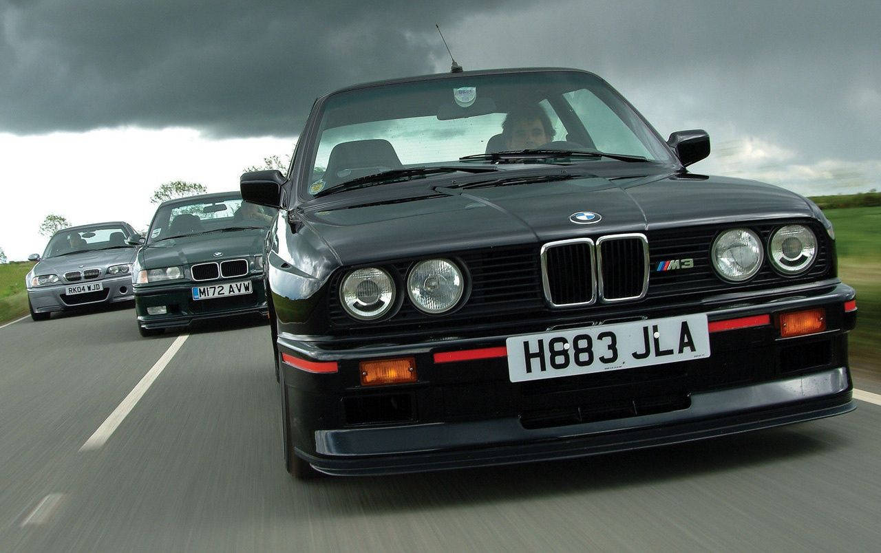BMW M3 系列賽車 壁紙 — 三輛黑色BMW M3跑車在暴風雨灰暗的天空下，在潮濕的道路上奔馳，呈現模糊的動態效果。