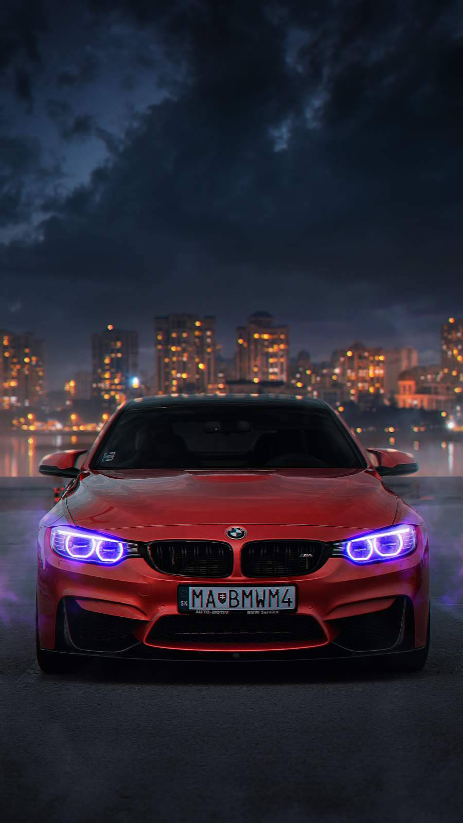 紅色BMW M4城市風景iPhone車壁紙 壁紙 — 一輛紅色寶馬跑車帶有發光的藍色車頭燈，在夜晚濕漉漉的城市街道上向前行駛，背景是黑暗多雲的天空和燈火通明的建築物。