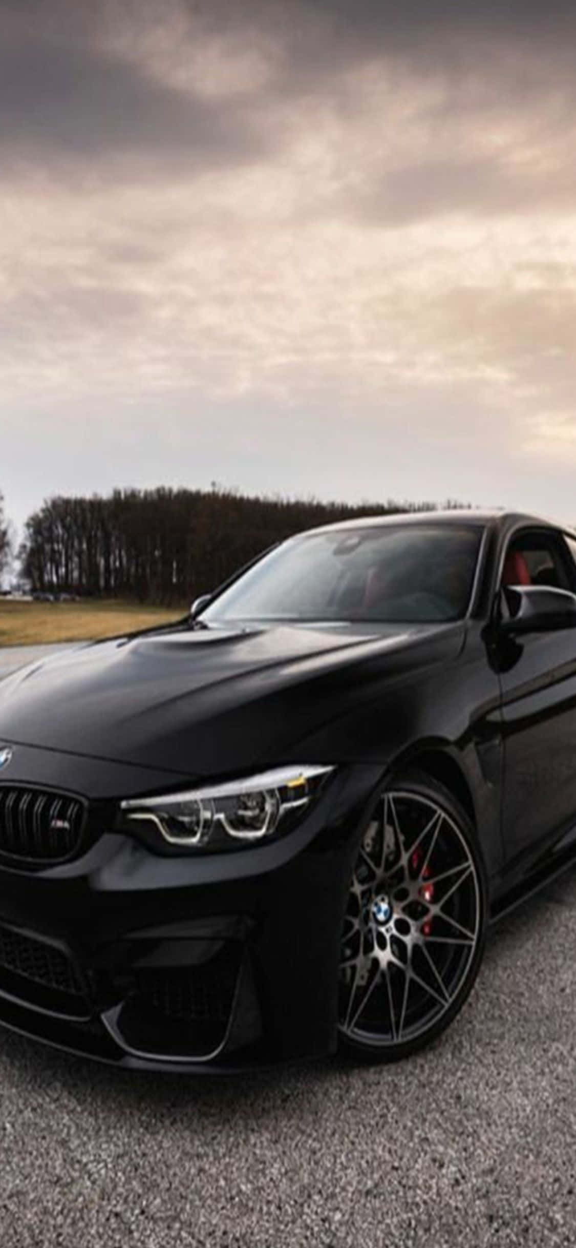 BMW M4 Coupe 與 iPhone X：完美搭配 壁紙 — 一輛時尚的黑色BMW M4跑車停在碎石上，陰沉多雲的天空下，遠處有樹木。