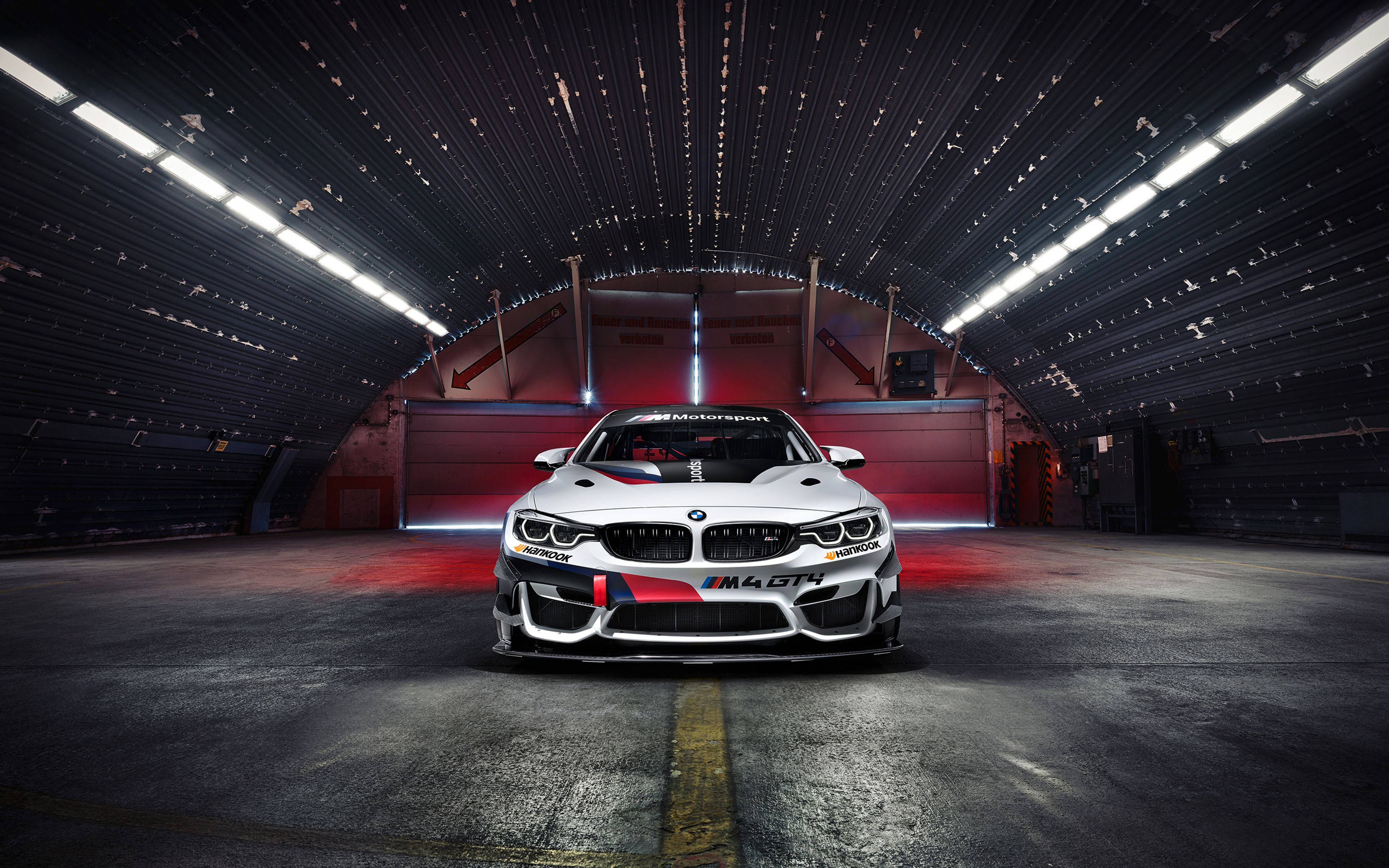 BMW M4 GT4 豪華跑車 壁紙 — 一輛白色BMW M4 GT4賽車帶有紅色點綴，在黑暗的工業車庫中，明亮的頂燈下居中停放。
