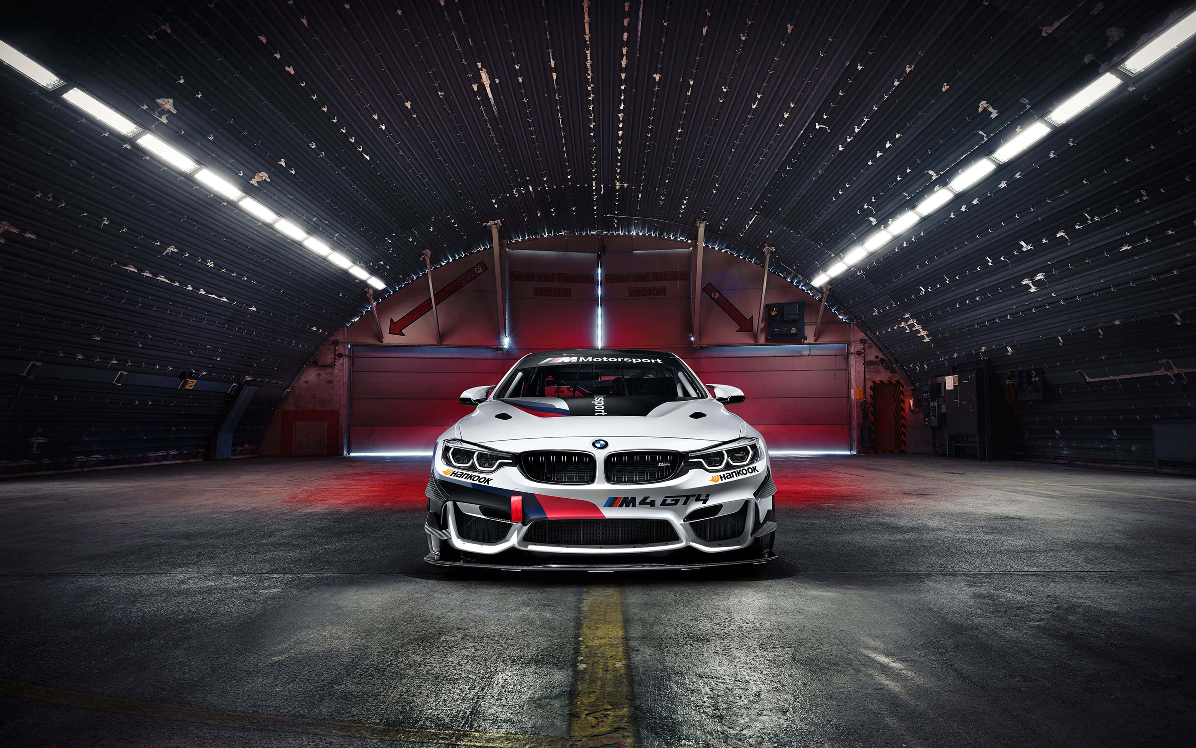 BMW M4 GTS客製化平板壁紙 壁紙 — 一輛帶有紅色點綴的白色BMW M4 GT4賽車，在明亮的頂燈下居中停放在黑暗的工業車庫中。