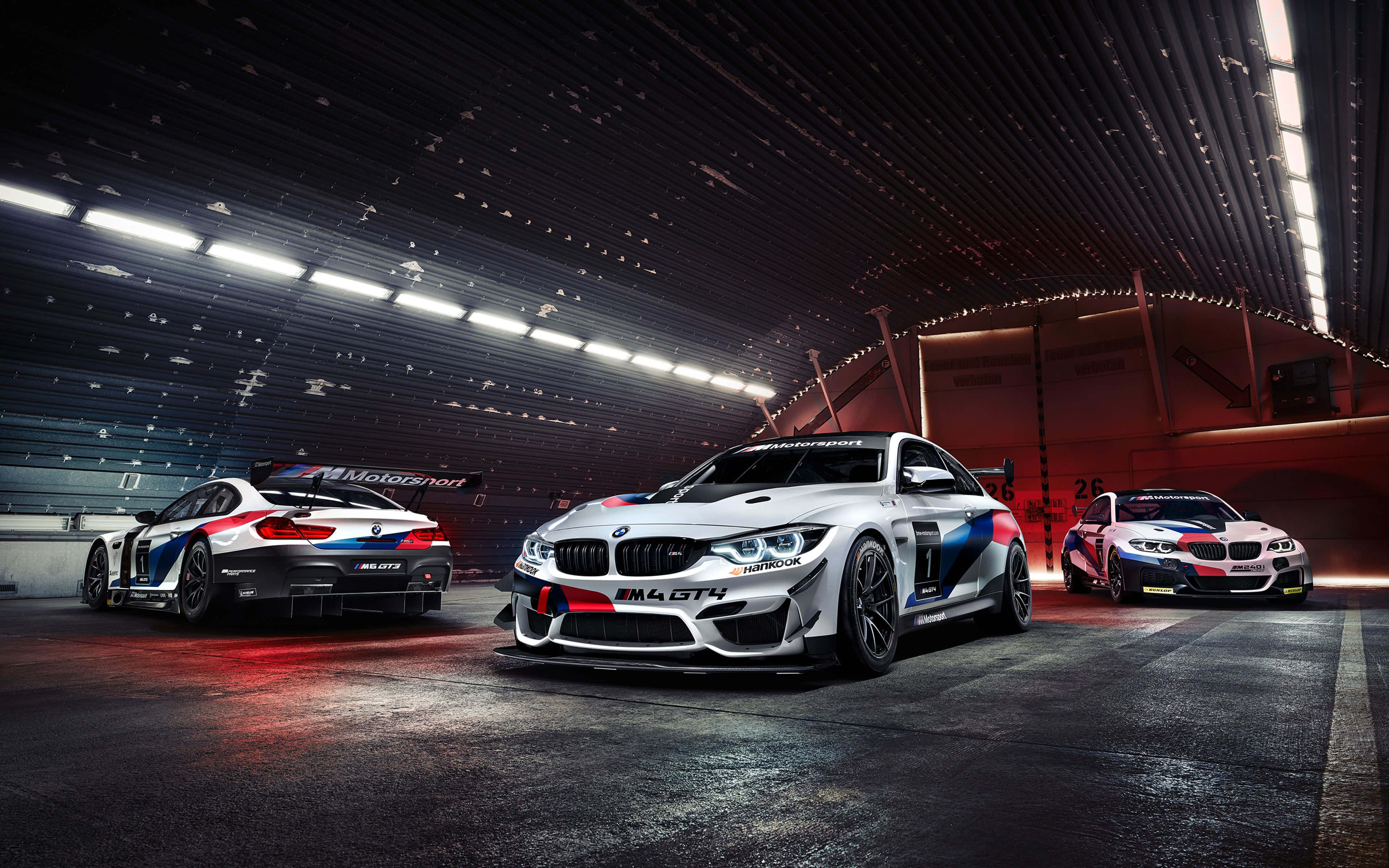 BMW M4 GTS 白漆紅藍彩繪背景 壁紙 — 三輛白色、藍色和紅色塗裝的BMW M4 GT4賽車停放在明亮的現代化車庫中，上方有明亮的頂燈。