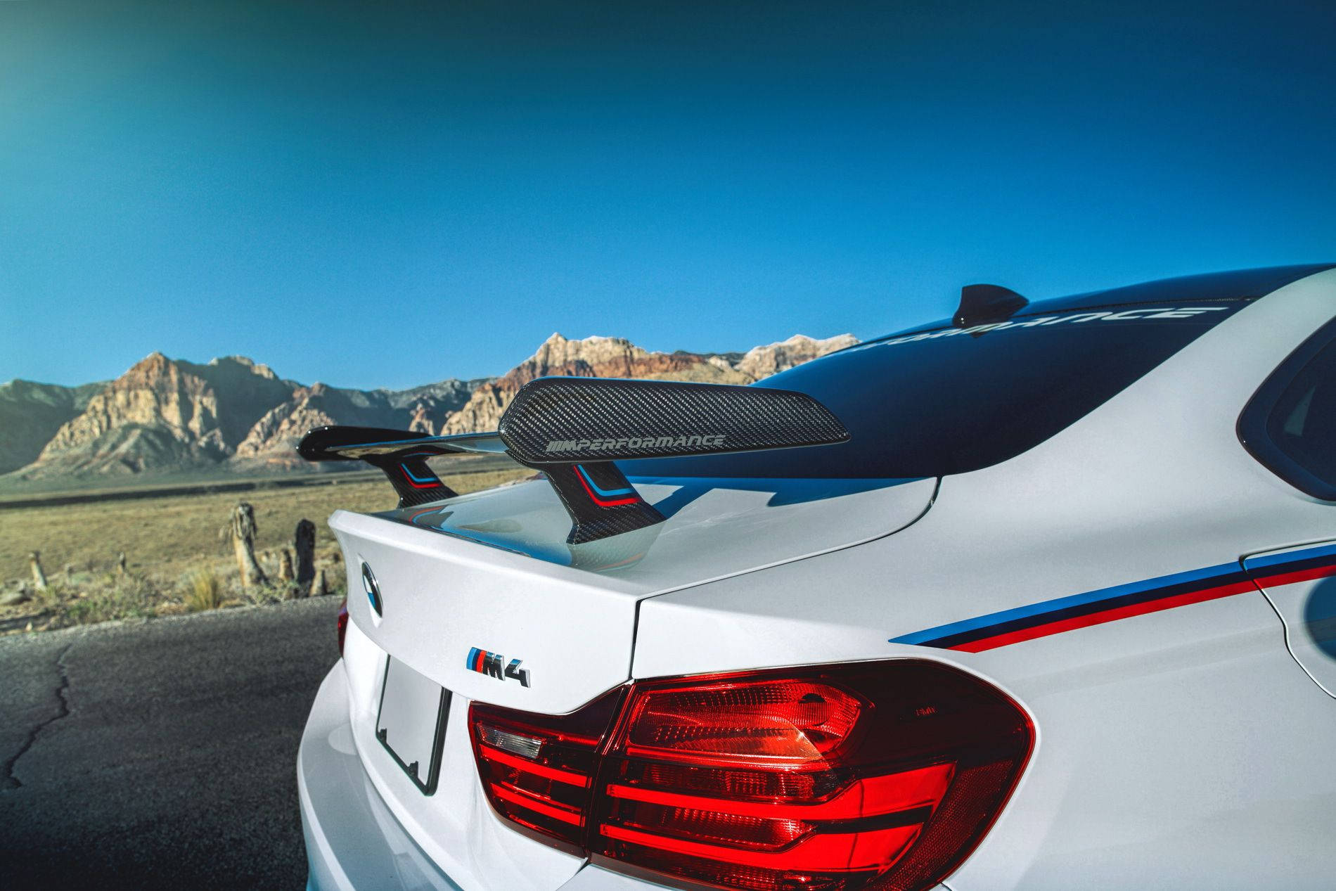 BMW M4 系列 壁紙 — 一輛白色BMW M4配備碳纖維尾翼，停泊在沙漠道路上，背景是岩石山脈和清澈的藍天。