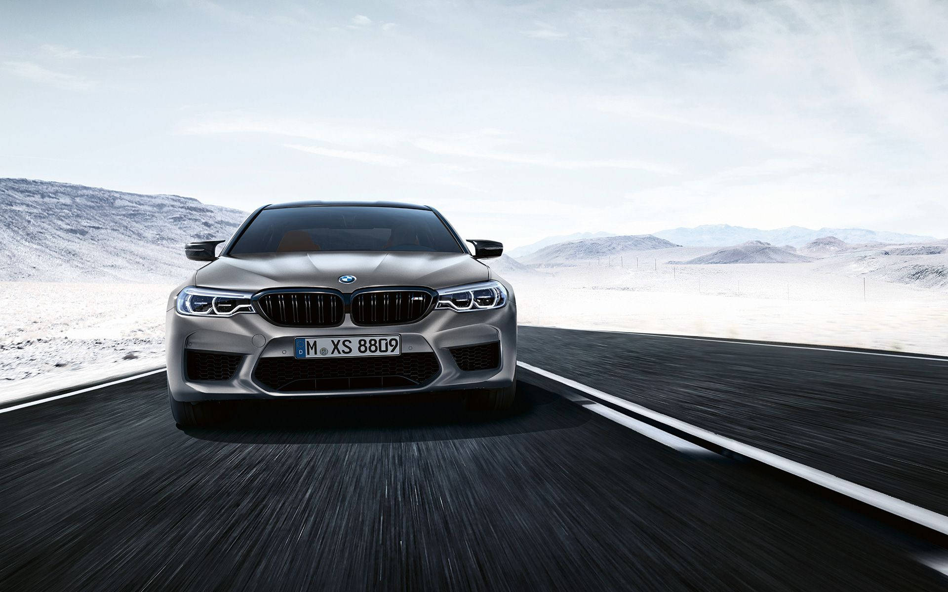 BMW M5系列跑車 壁紙 — 一輛銀色寶馬M5在沙漠高速公路上飛馳，遠方有白雪覆蓋的山脈，天空明亮。
