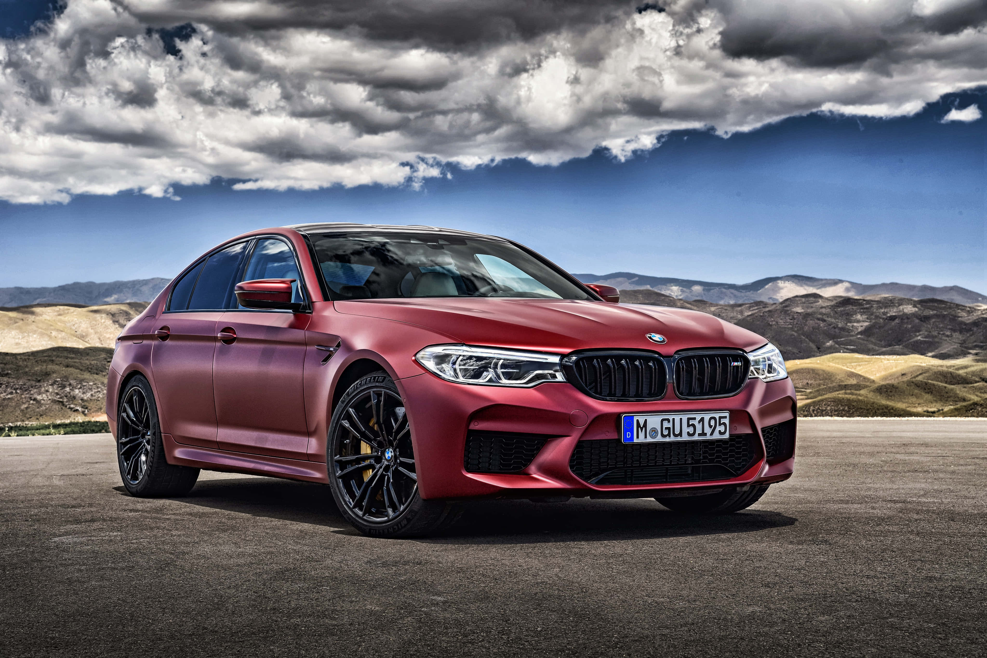 體驗 BMW M5 的奢華動力 壁紙 — 一輛啞光紅色的BMW M5運動轎車停在瀝青路上，背景有戲劇性的雲層和山區地形。