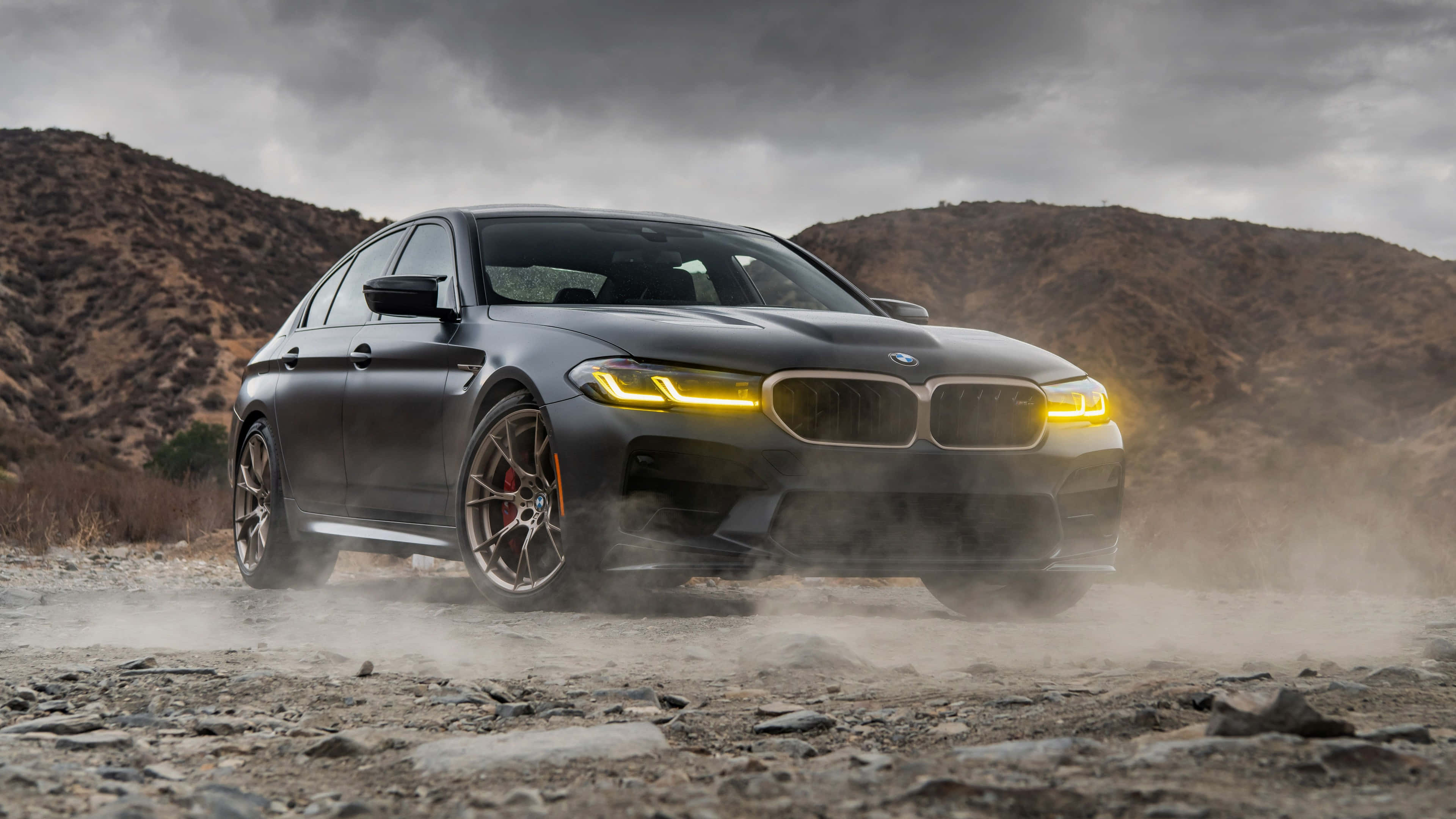 BMW M5 4K - 強勁動感的完美化身 壁紙 — 一輛啞光黑色BMW M5在塵土飛揚的地形中飄移，黃色車頭燈在暴風雨的天空下閃耀。