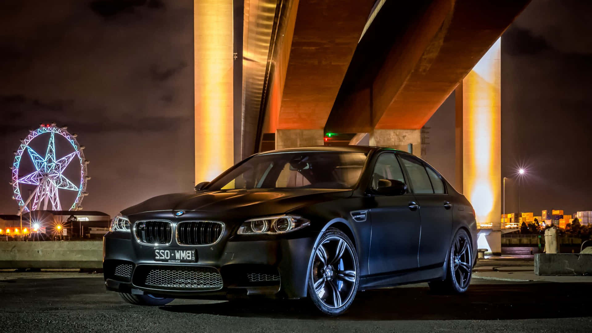啞光黑色BMW M5在夜間停泊在亮燈的橋下，靠近摩天輪。