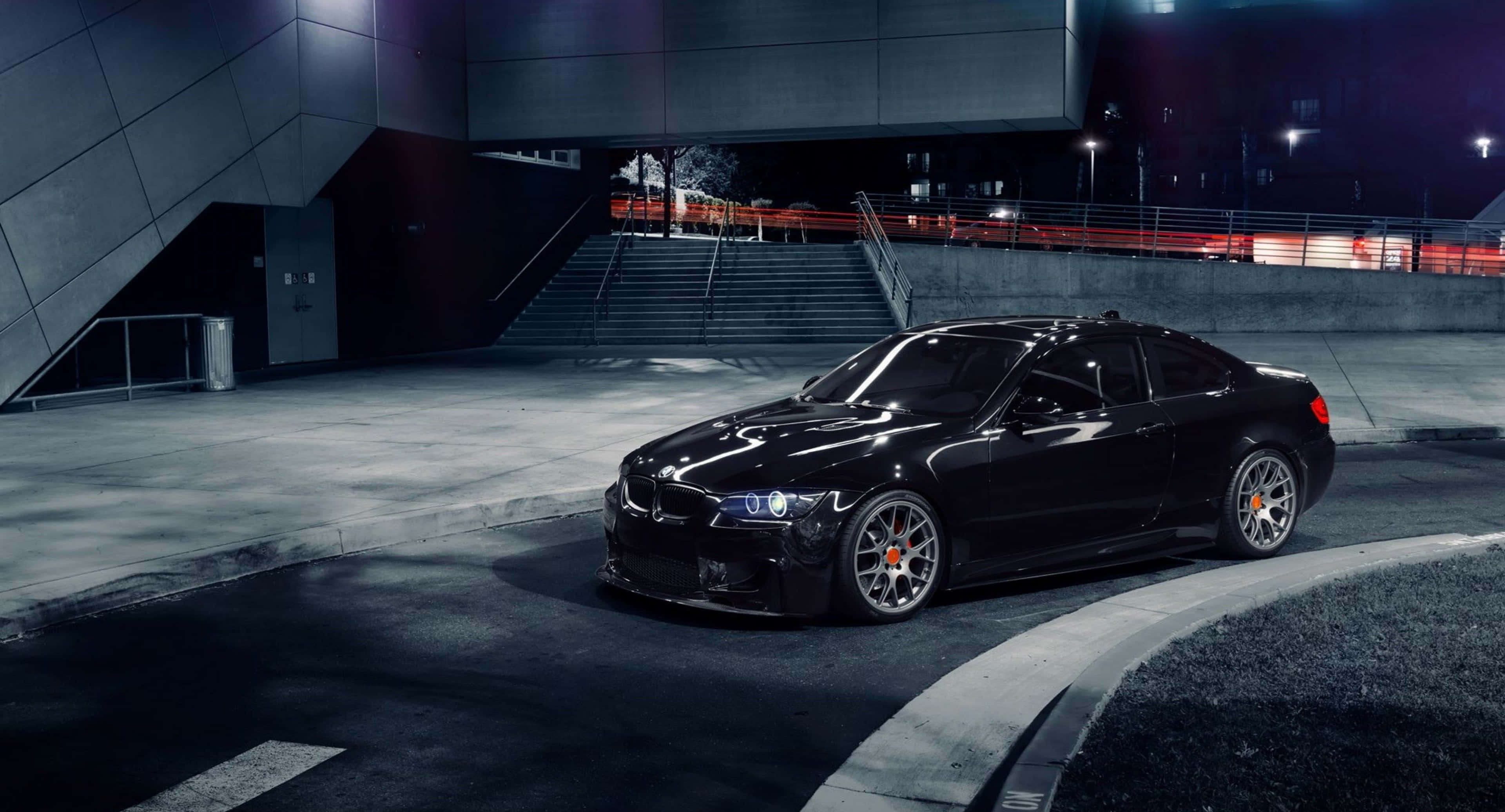 BMW M5路面上漂移平板壁紙 壁紙 — 一輛時尚的黑色跑車在人造燈光下閃閃發光，停泊在現代化的城市廣場上，夜色中。