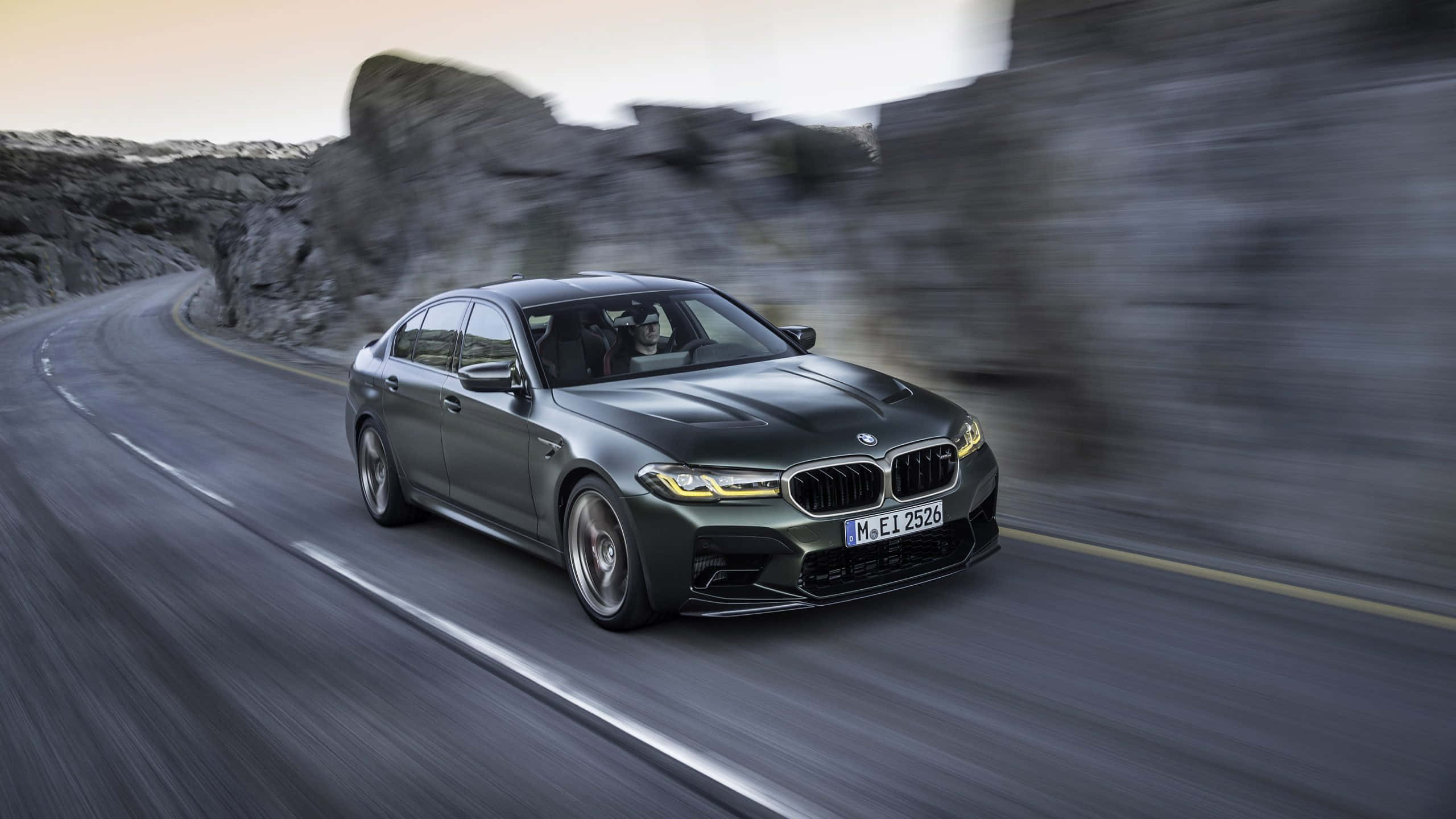 體驗 BMW M5 的澎湃動力 壁紙 — 一輛啞光灰色BMW M5沿著蜿蜒的山路疾馳，伴隨著模糊的岩石懸崖和瀝青動態模糊。