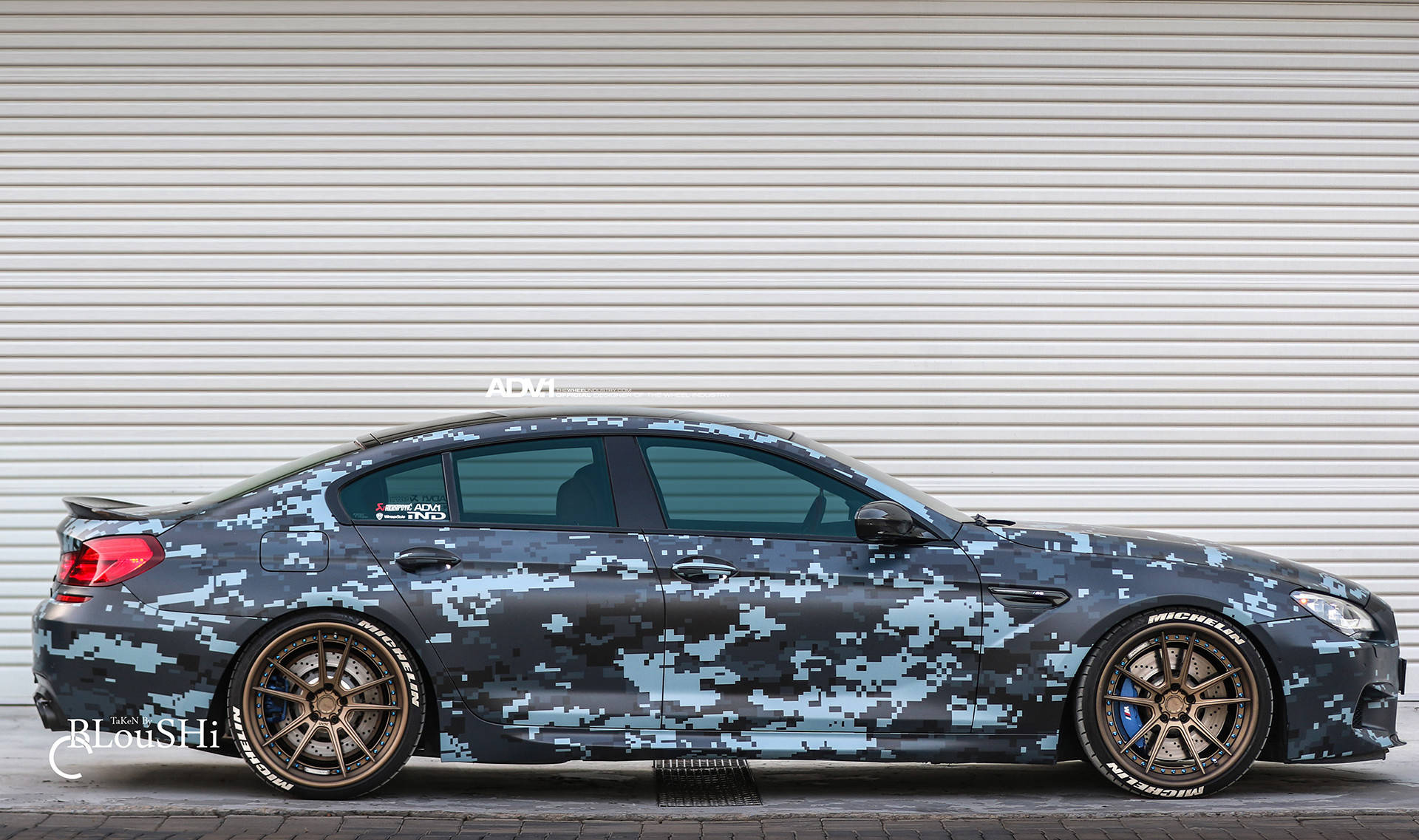 黑色迷彩BMW M5展現隱蔽奢華 壁紙 — 一輛時尚的BMW M5跑車，帶有數碼迷彩包膜，停靠在波紋金屬車庫門前。