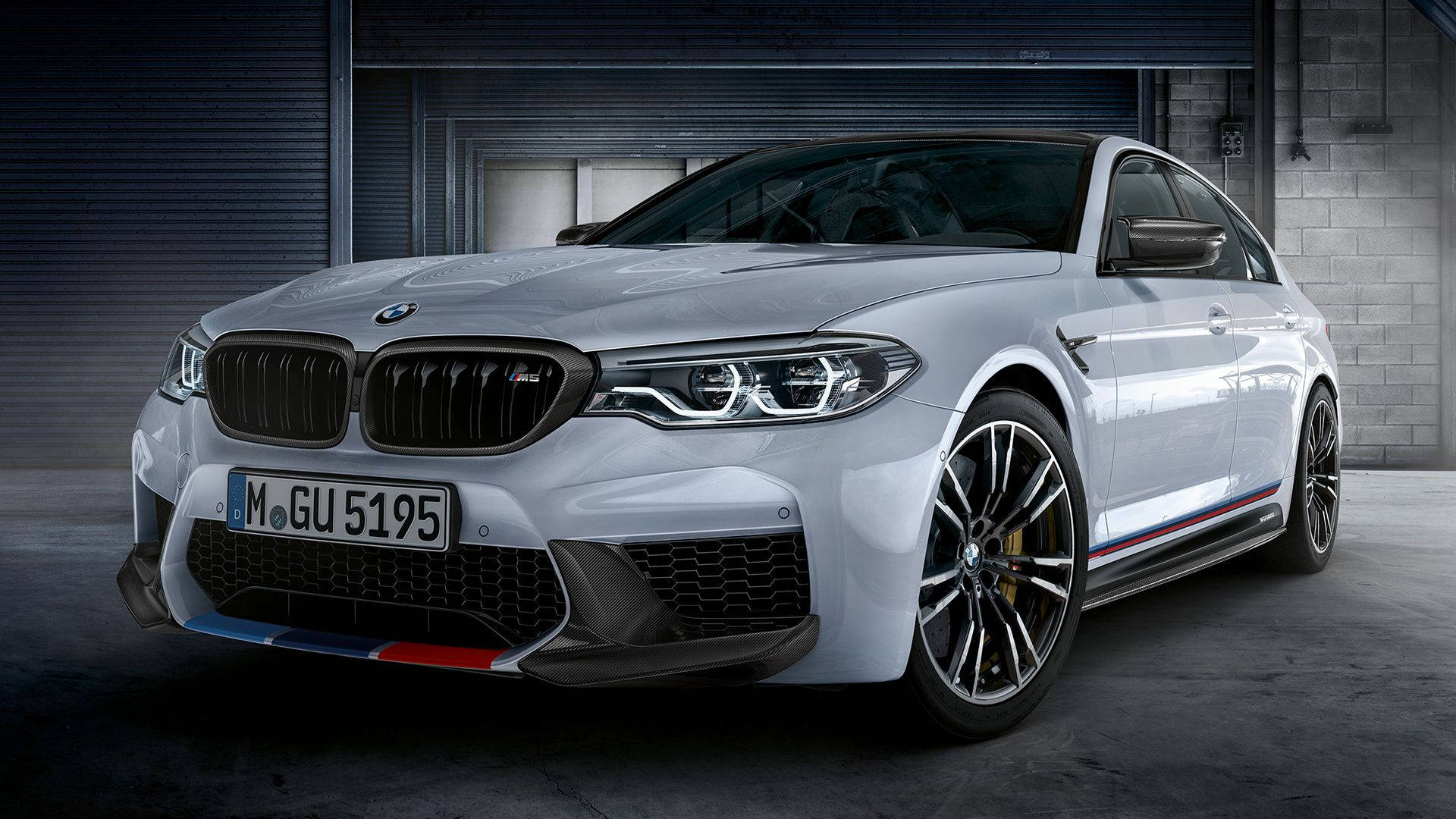 BMW M5 系列白色款 壁紙 — 一輛時尚的銀色BMW M5轎車，帶有紅、藍、白賽車條紋，停放在光線昏暗的工業車庫中。