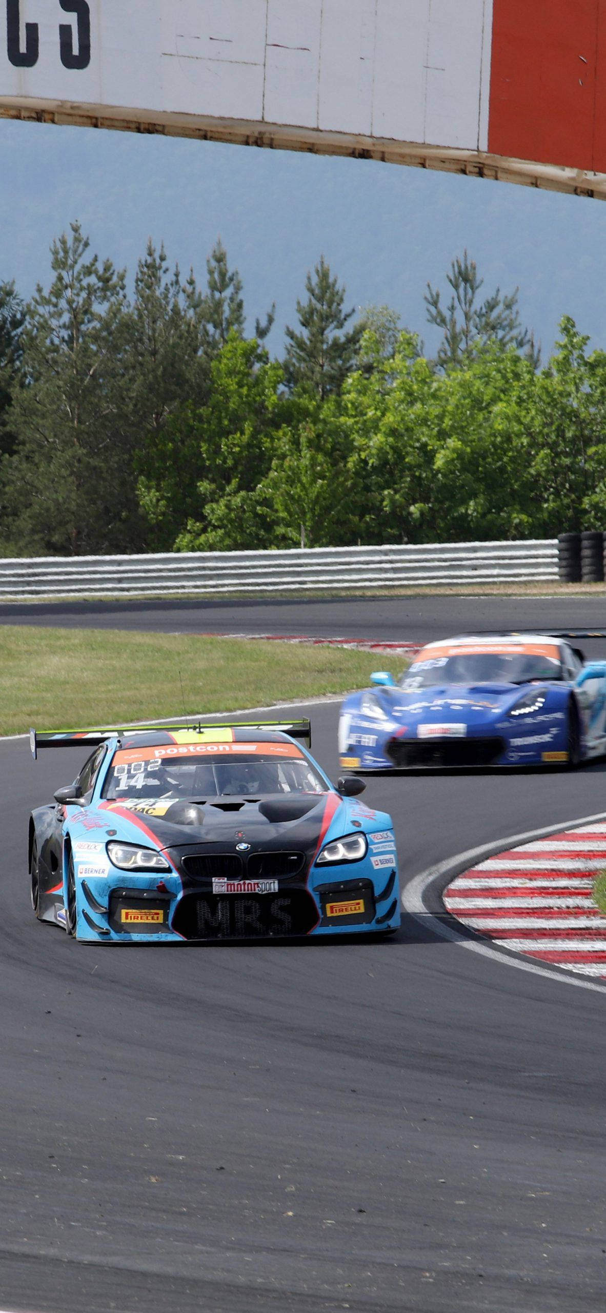 藍色 BMW M6 GT3 賽車 iPhone 壁紙 — 兩輛賽車在陽光明媚的瀝青賽道上高速駛過一個急彎，賽道兩旁綠樹成蔭，並有紅白相間的賽道邊緣。