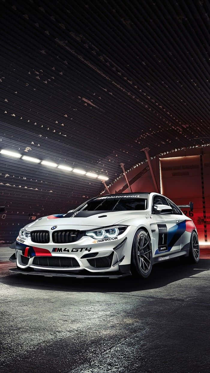 你駕駛的不只是任何車，而是 BMW M8 壁紙 — 一輛帶有藍紅飾邊的白色BMW M4 GT4賽車，在明亮的燈光下，停放在時尚的暗色車庫中。