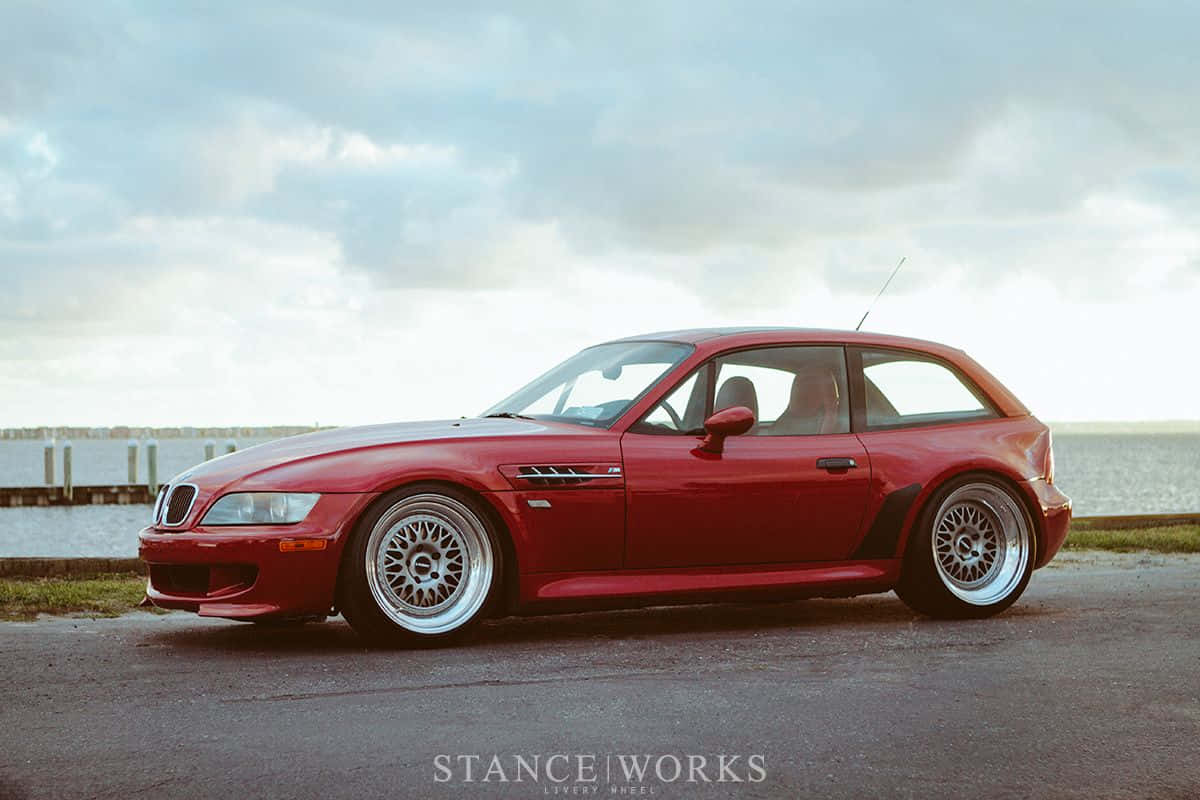 BMW M8 Gran Coupé概念車 壁紙 — 一輛光亮的紅色BMW Z3 M Coupe配備定制輪圈，停泊在多雲天空下的水邊瀝青路上。