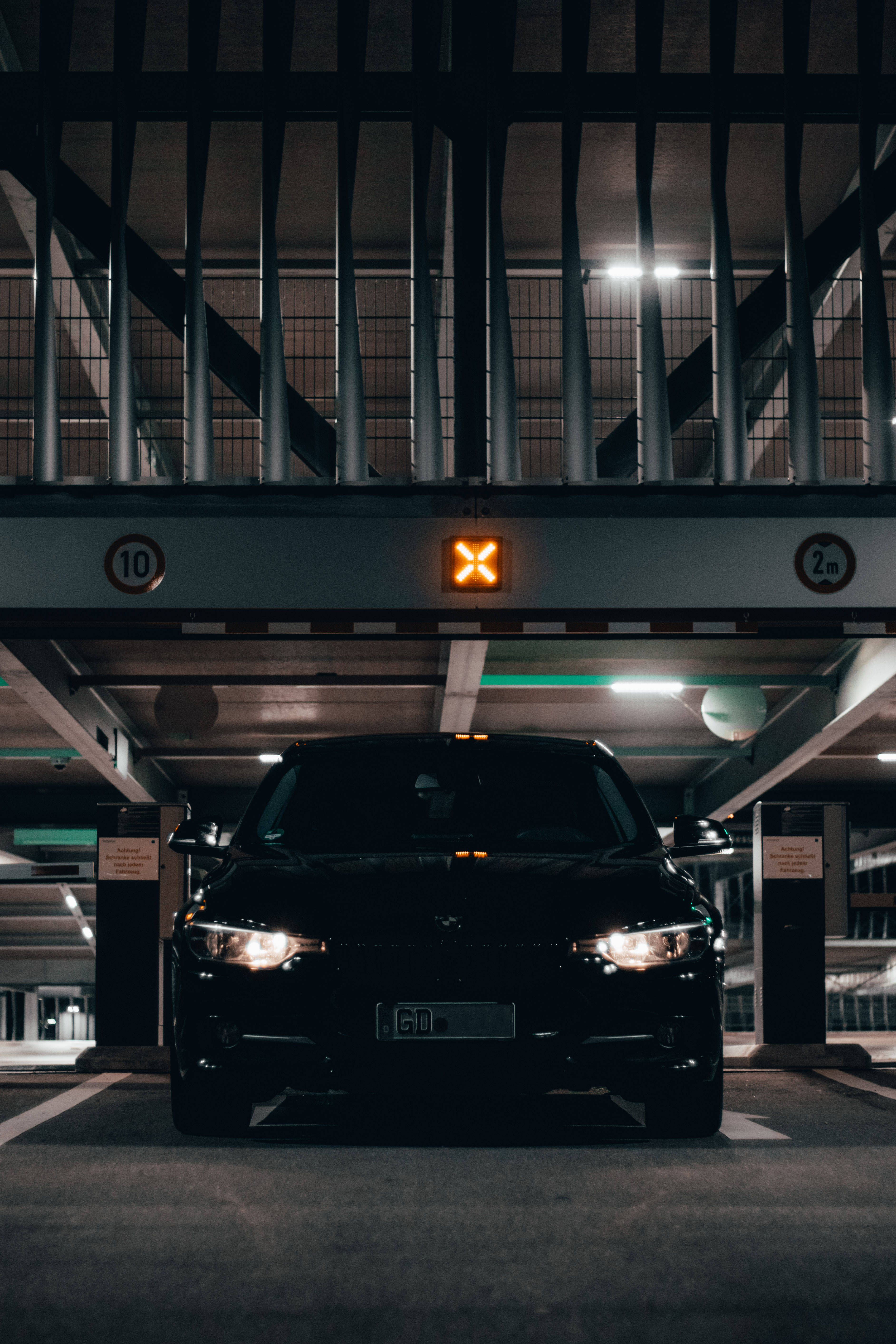 BMW M系列停泊於停車場 壁紙 — 一輛時尚的黑色汽車在燈光昏暗的停車場內向前，上方有一個發光的橙色「X」標誌。