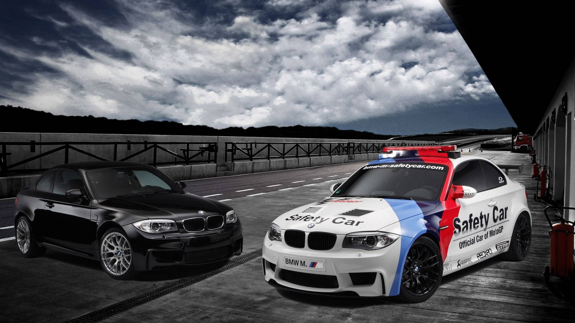 BMW M系列安全車 壁紙 — 兩輛寶馬賽車，一黑一白的安全車帶有紅藍條紋，停在賽道上，背景是戲劇性的暴風雲。