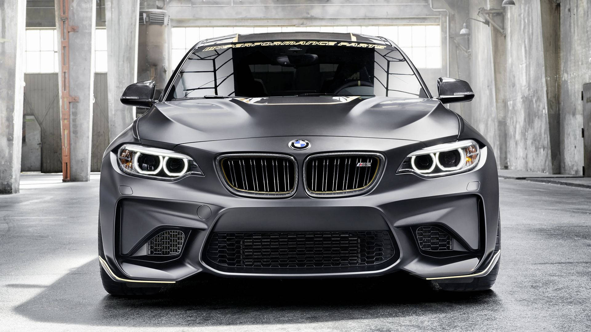 BMW M系列攝影棚拍攝 壁紙 — 一輛啞光灰色BMW M2跑車帶金色飾邊停放在混凝土工業倉庫內。