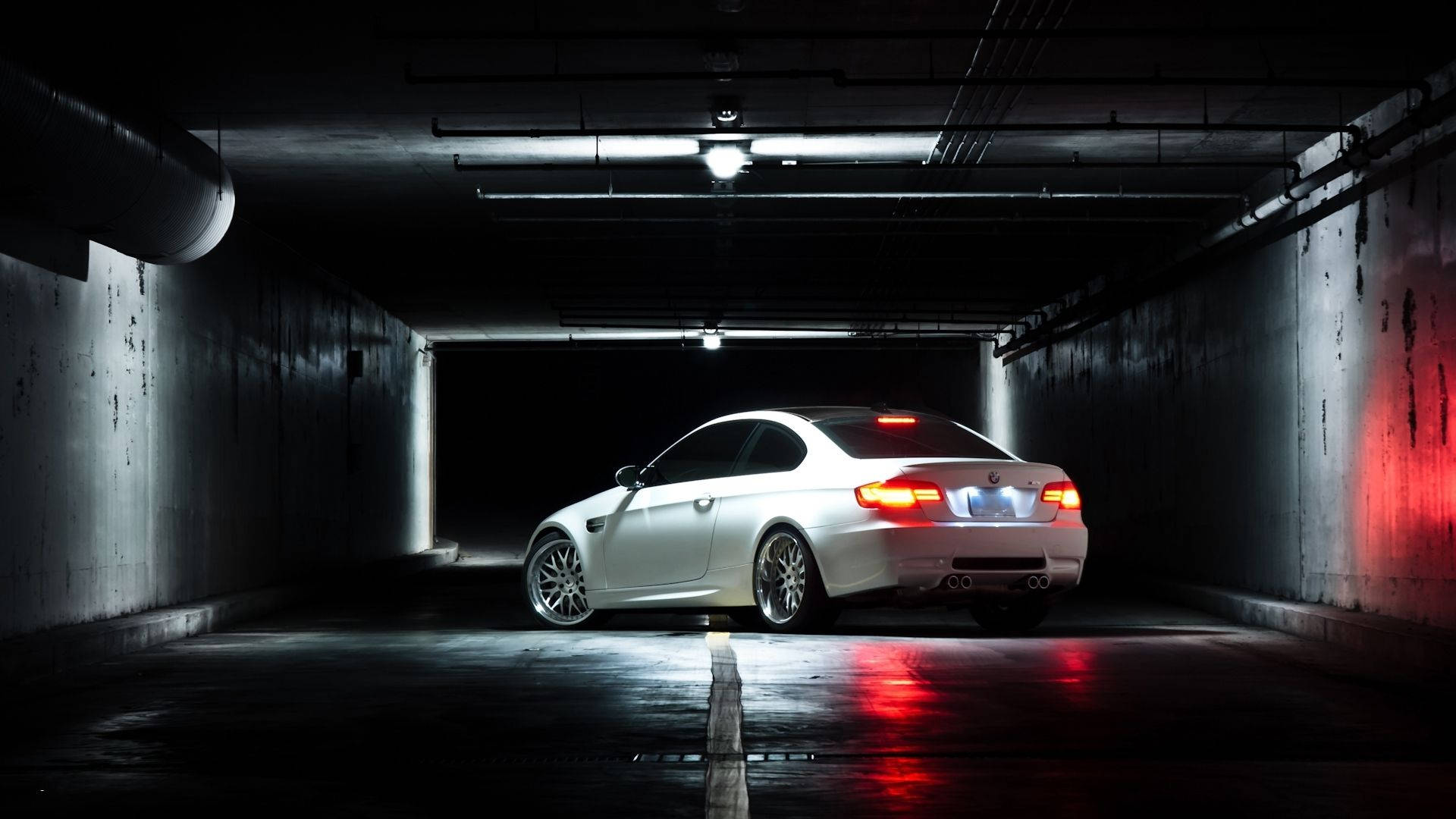 BMW M系列跑車暗色車庫 壁紙 — 一架時尚的白色跑車，紅色尾燈發光，停泊在光線昏暗、混凝土地面潮濕的地下停車場。