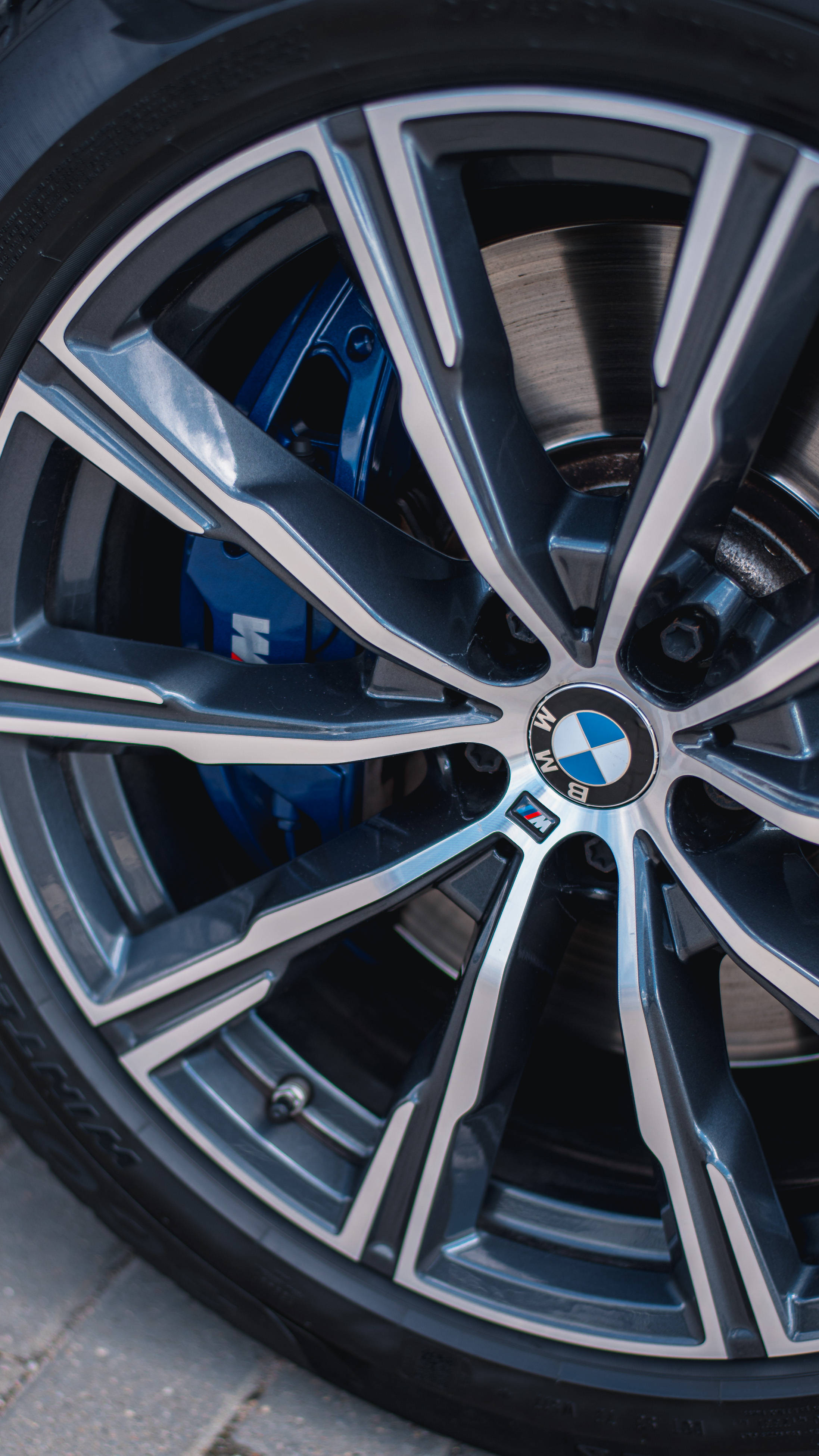 BMW M車輪與車標特寫 壁紙 — BMW M 型號車輪特寫，展示銀色輪輻、藍色制動卡鉗和路面上的輪胎花紋。