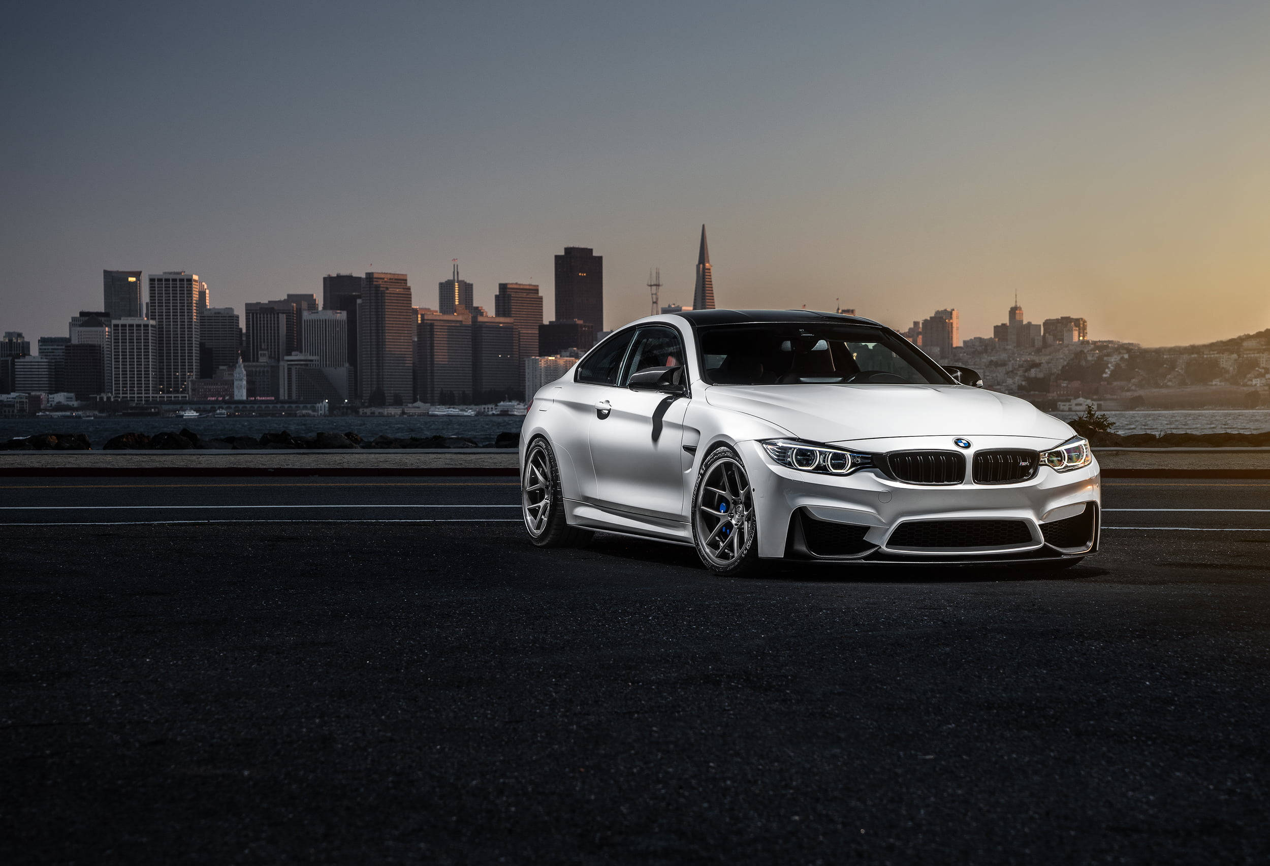 簡約白色 BMW 電腦 壁紙 — 一架時尚的銀色BMW M4雙門跑車停泊在黑暗的道路上，夕陽下城市天際線閃耀。