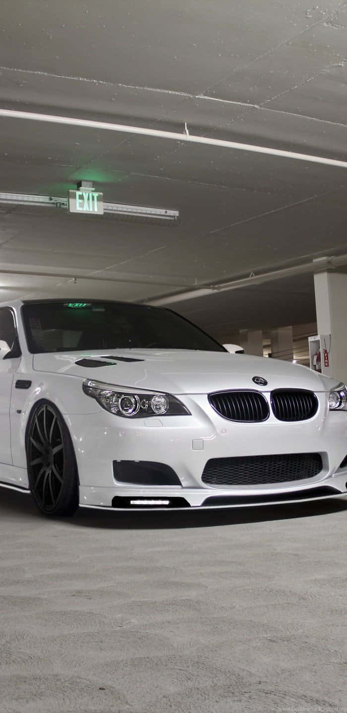 適用於 BMW 的 Porsche 充電系統 壁紙 — 一輛時尚的白色BMW M5跑車配黑色輪圈，停泊在昏暗的地下停車場，上方有綠色出口標誌。