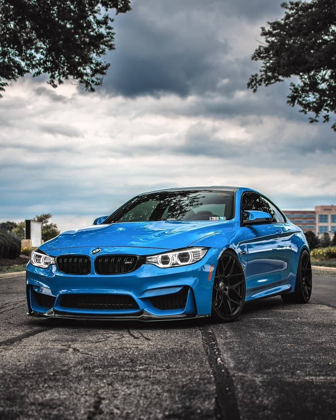 藍色BMW停泊於樹木旁 壁紙 — 一輛充滿活力的藍色BMW M4轎跑車在樹枝框住的戲劇性多雲天空下閃閃發光。