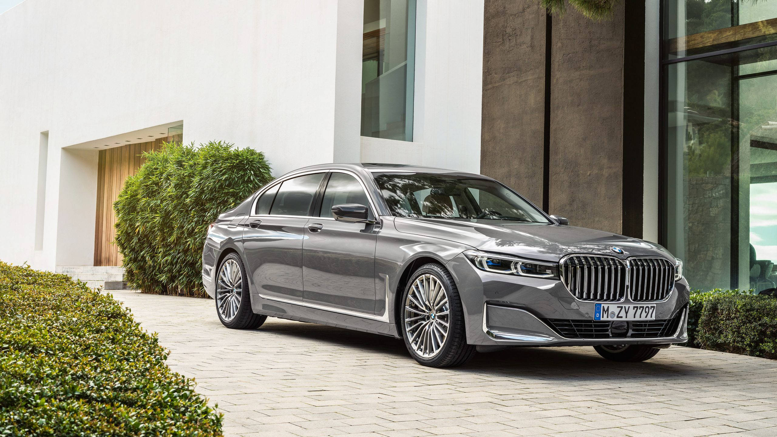BMW科技之美 壁紙 — 一架時尚的銀色寶馬7系轎車停泊在石頭車道上，旁邊是現代建築和修剪整齊的綠色樹籬。