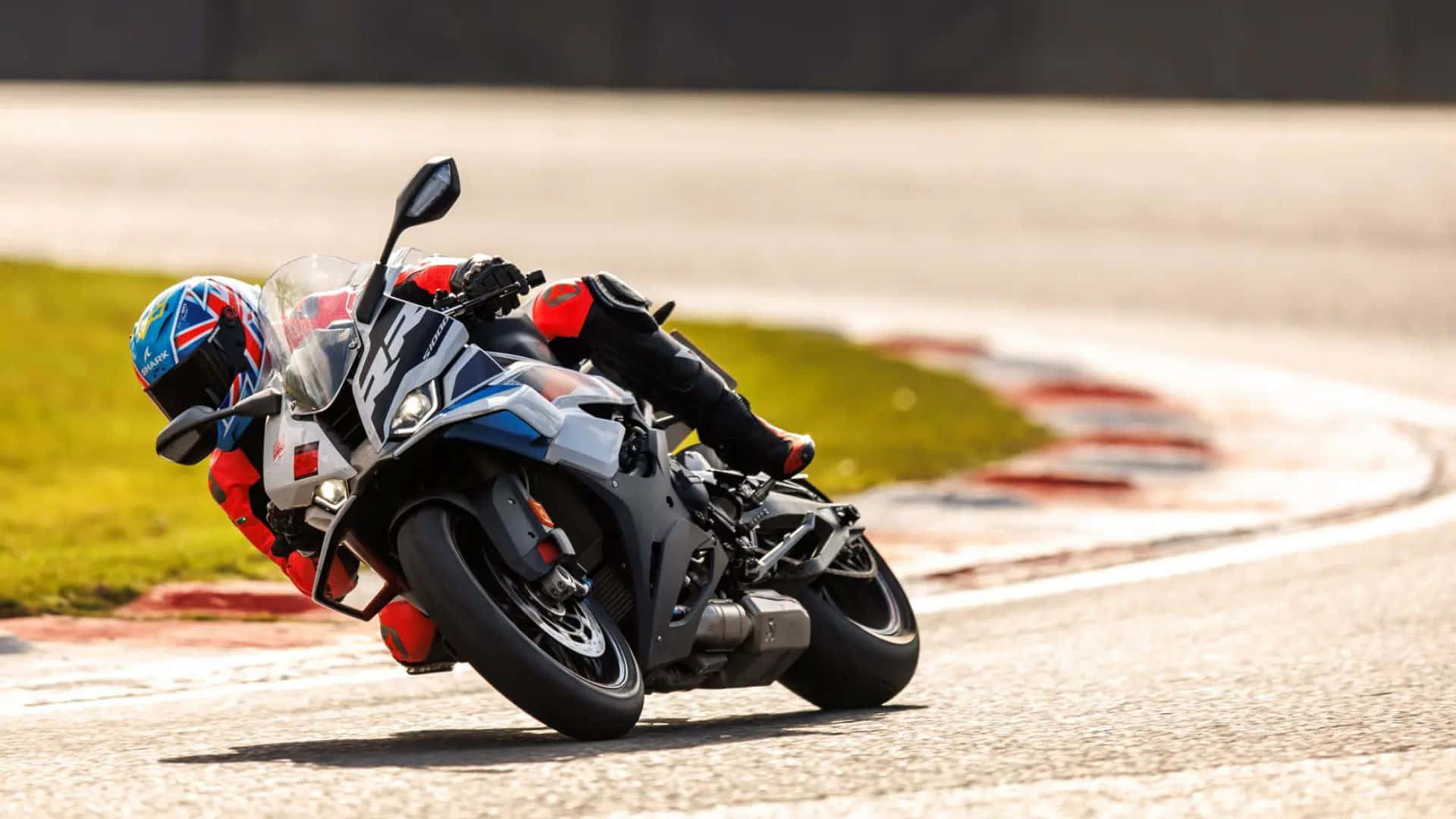 BMW S1000RR 賽車競速動作 壁紙 — 一名騎手在陽光普照的賽道上，騎著運動型摩托車傾斜入一個急彎，賽道有紅色和白色的路緣石。