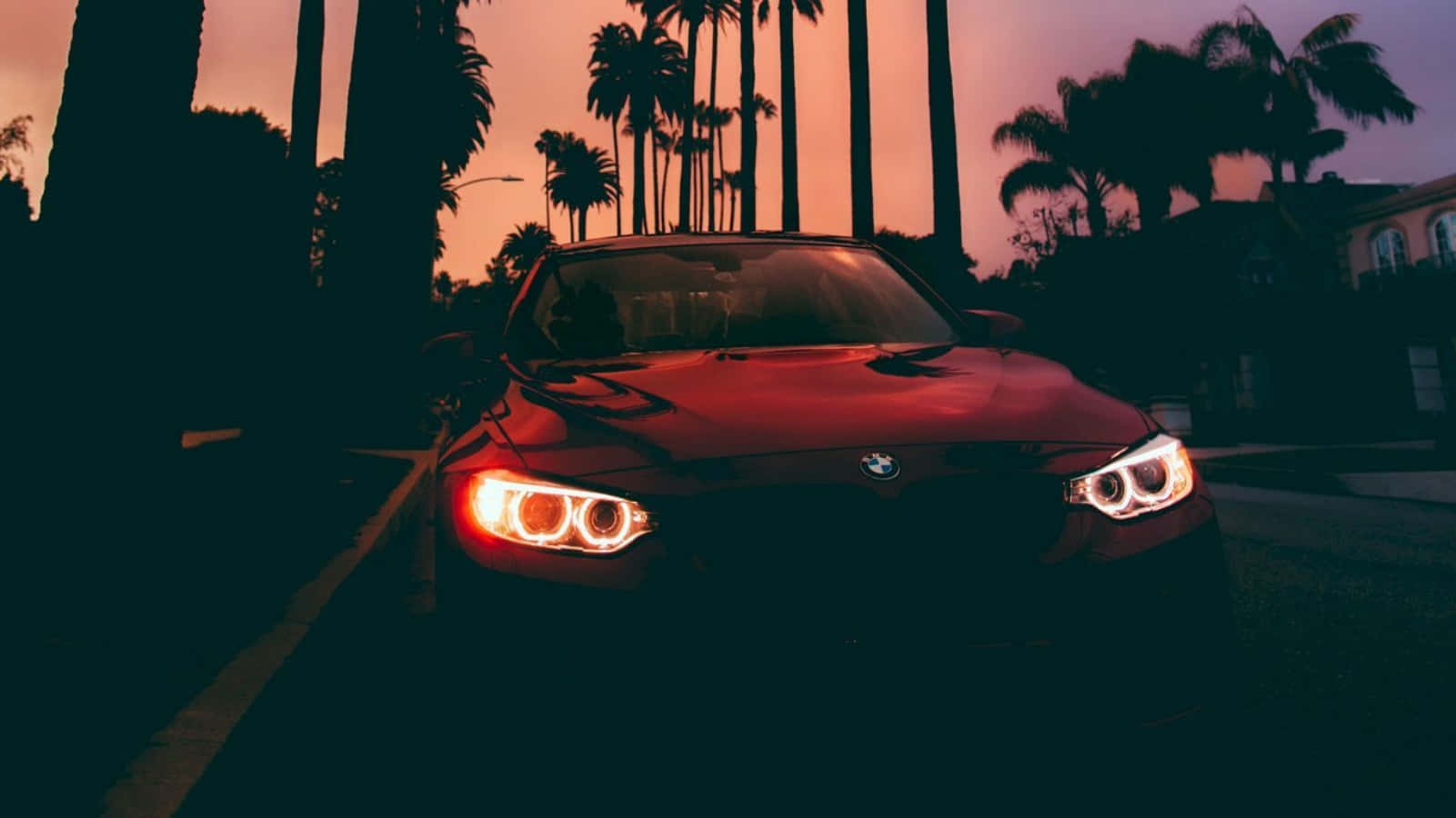 BMW 美學標誌與黃色頭燈夜景平板壁紙 壁紙 — 一輛紅色寶馬跑車在暮色天空下閃爍著車頭燈，背景是高聳的棕櫚樹。